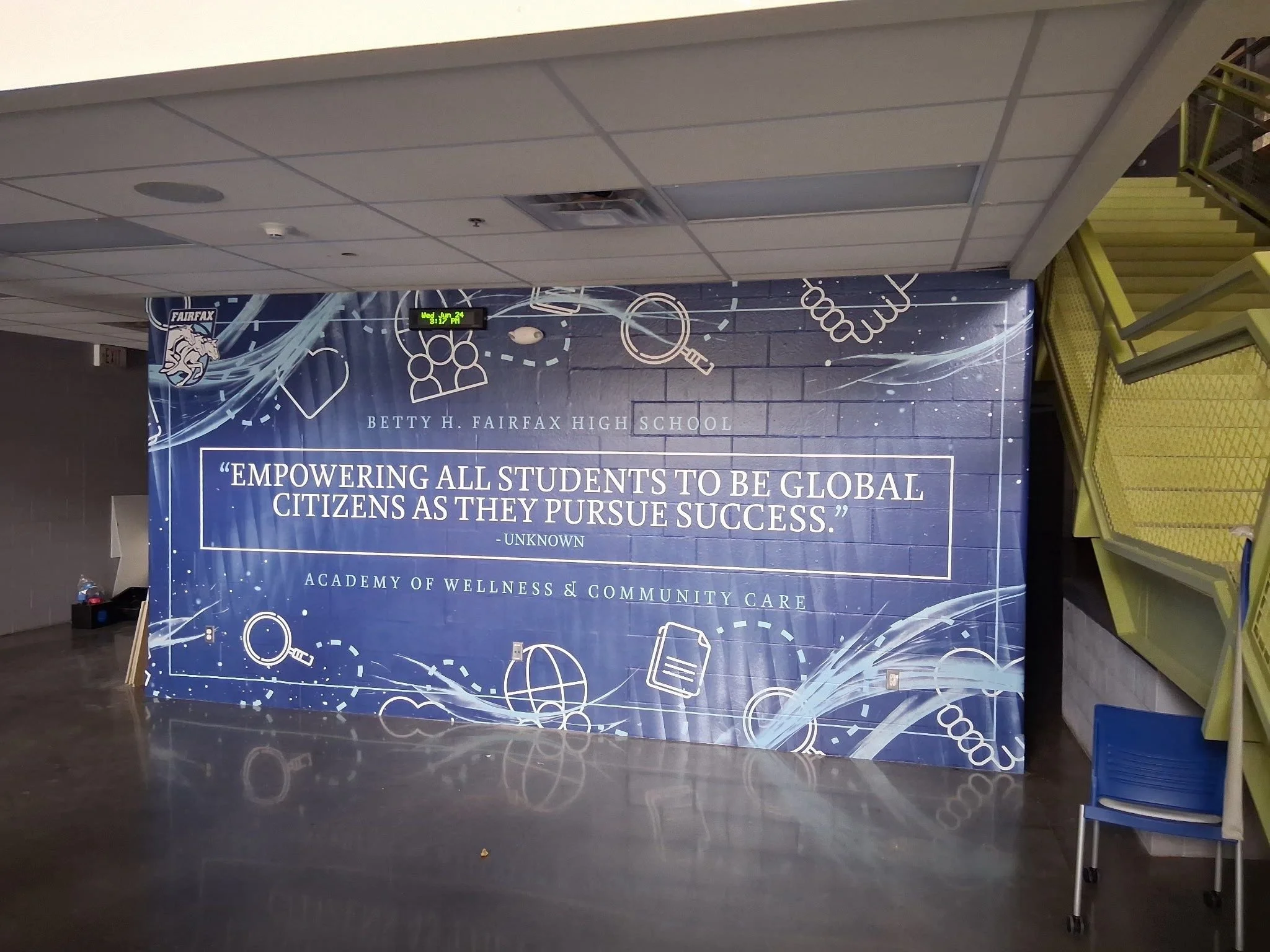 Indoor hallway with a large blue poster for Betty H. Fairfax High School, featuring science-themed icons and a quote about empowering students to be global citizens. Yellow seating is visible on the right.