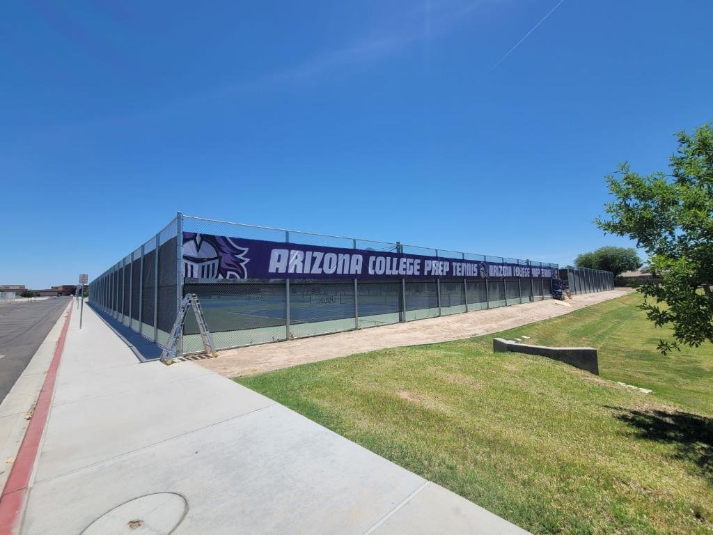 A fenced outdoor tennis court with a sign for Arizona College Prep Tennis, located on a grassy area under a clear blue sky.