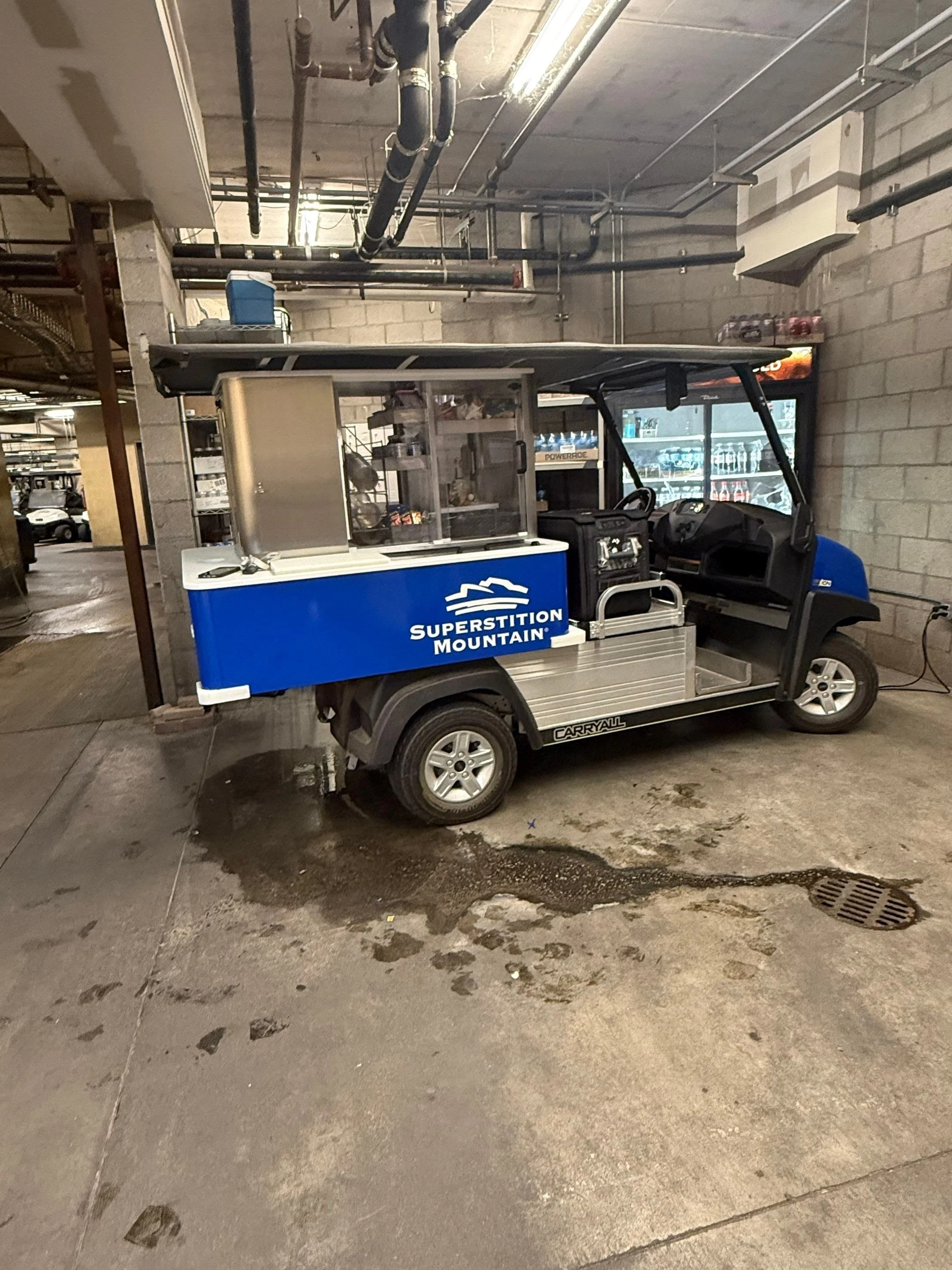 Electric golf cart with a blue and white color scheme, labeled 'Superstition Mountain,' parked in an indoor garage with concrete floors and cinder block walls, with various pipes and a vending machine in the background.