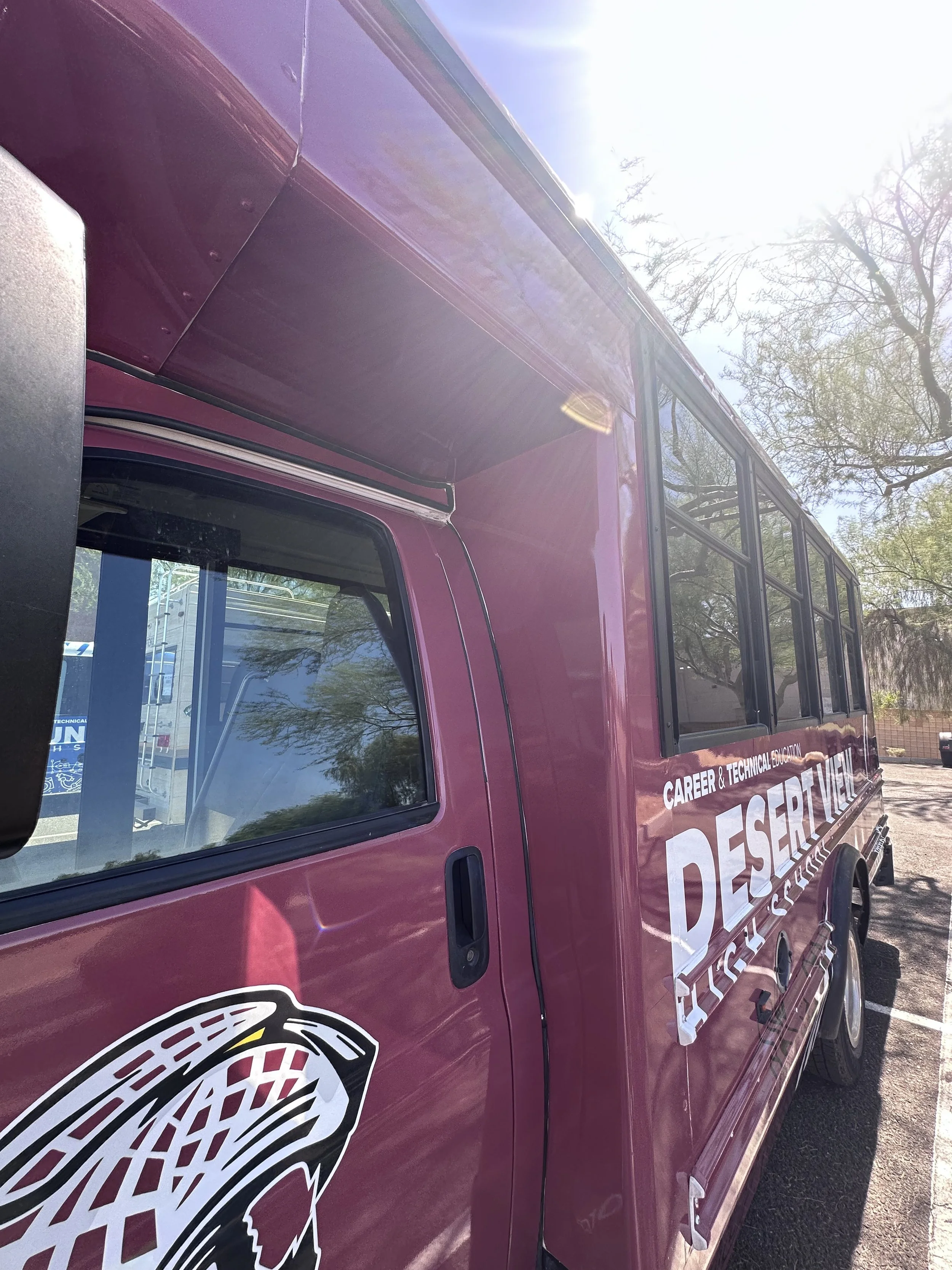 A maroon school bus with the words "Career & Technical Education Desert View" printed on it, parked outdoors under a tree, with the sun shining brightly overhead.