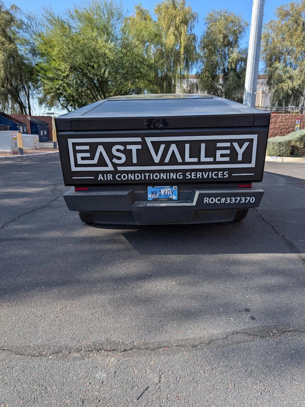 Rear view of a vehicle with a sign reading "East Valley Air Conditioning Services" and a Arizona license plate, parked on a paved lot with trees and trash bins in the background.