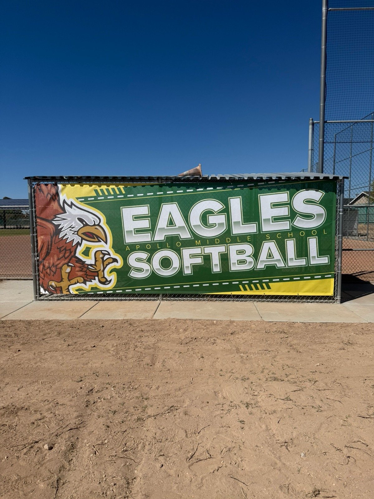 Green banner with a lion mascot and the text "Eagles Softball, Apollo Middle School" on a fence at a softball field, with clear blue sky and part of the field visible in the background.