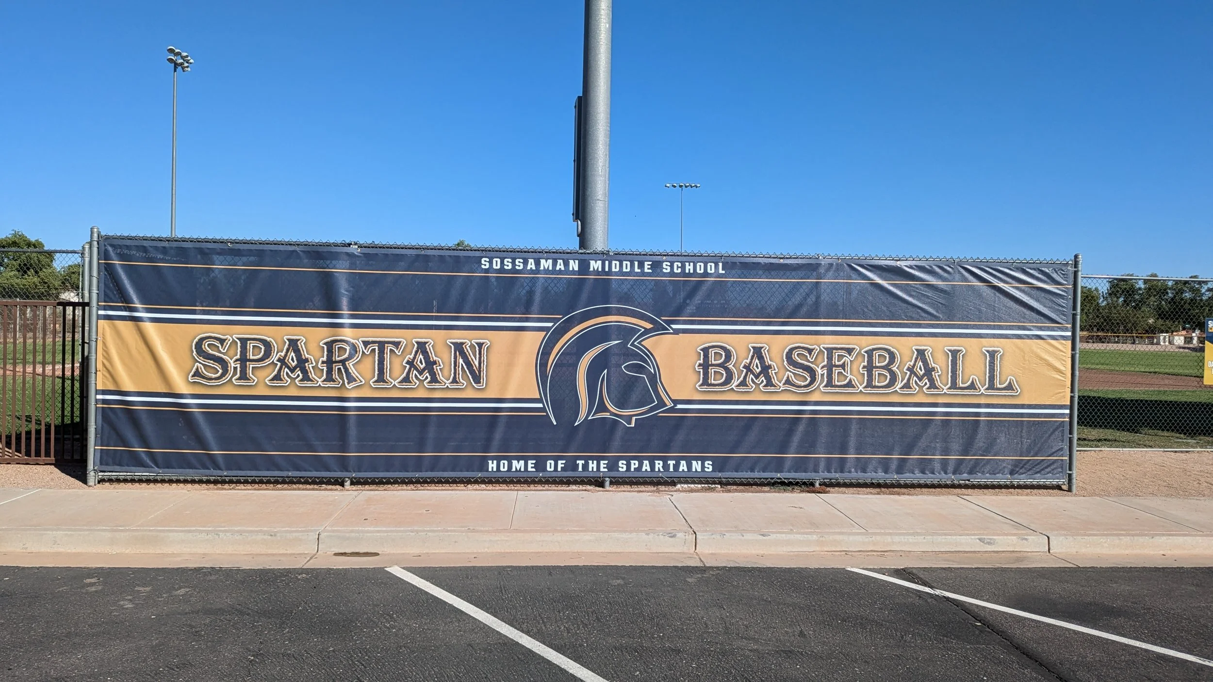 A large banner on a chain-link fence at an outdoor baseball field. The banner displays the text 'Sossaman Middle School Spartans Baseball, Home of the Spartans' with a Spartan helmet logo in the center. The sky is clear and blue, with stadium lights visible in the background.