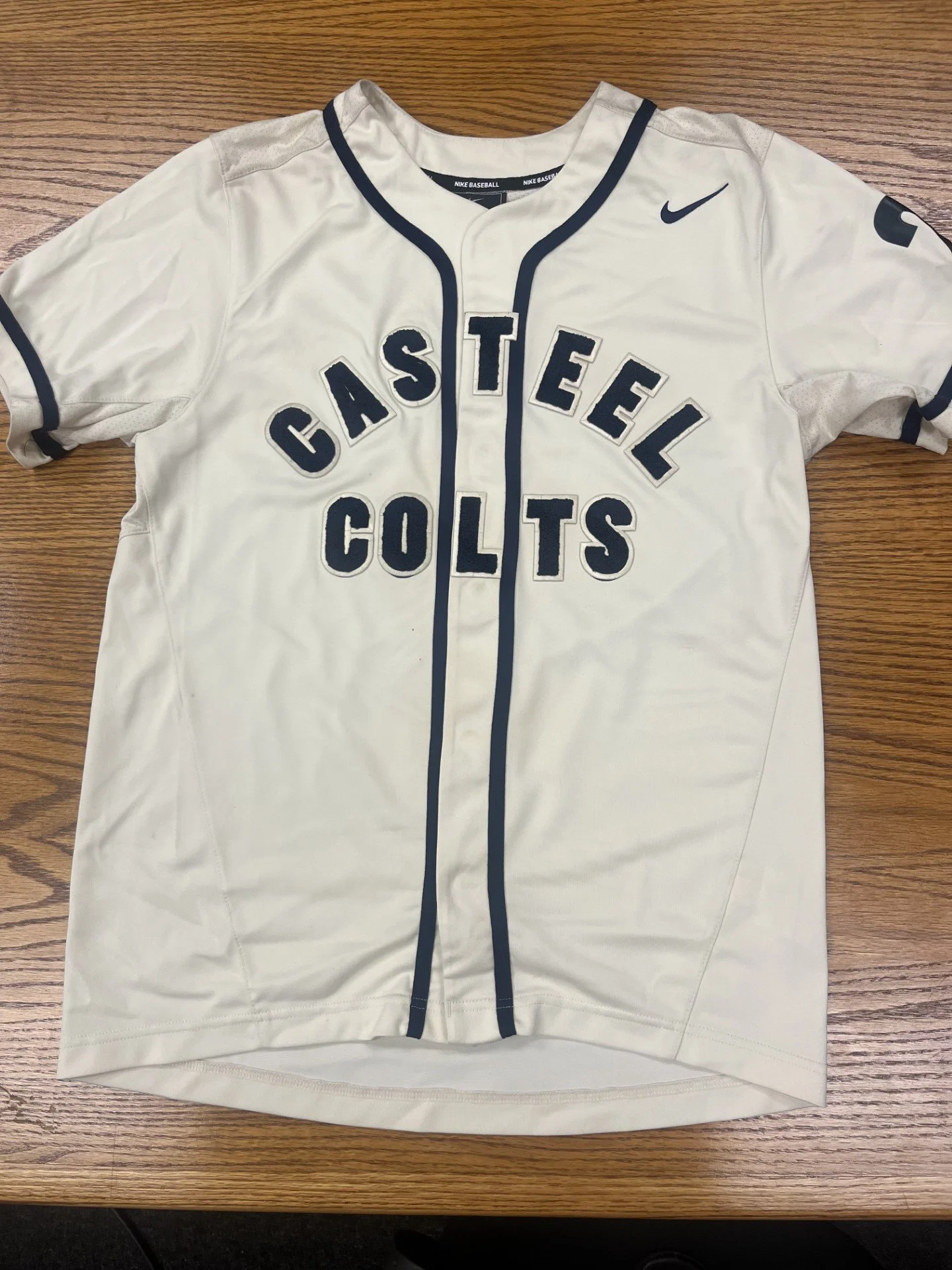 White sports jersey with black trim and the words "CASTEEL COLTS" printed on front, laid flat on a wooden surface.