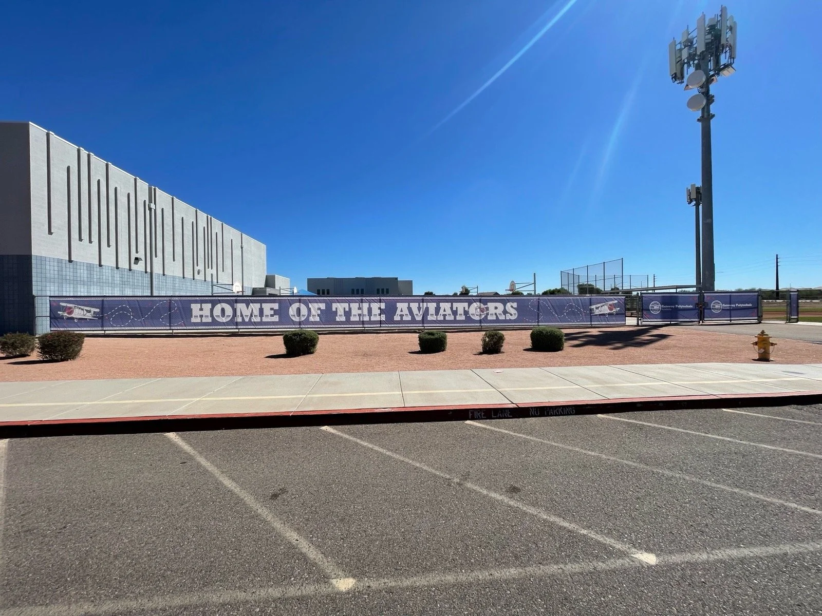 A billboard in a parking lot that reads 'Home of the Aviators' with an airplane graphic, behind a fence near a school building, under a clear blue sky.
