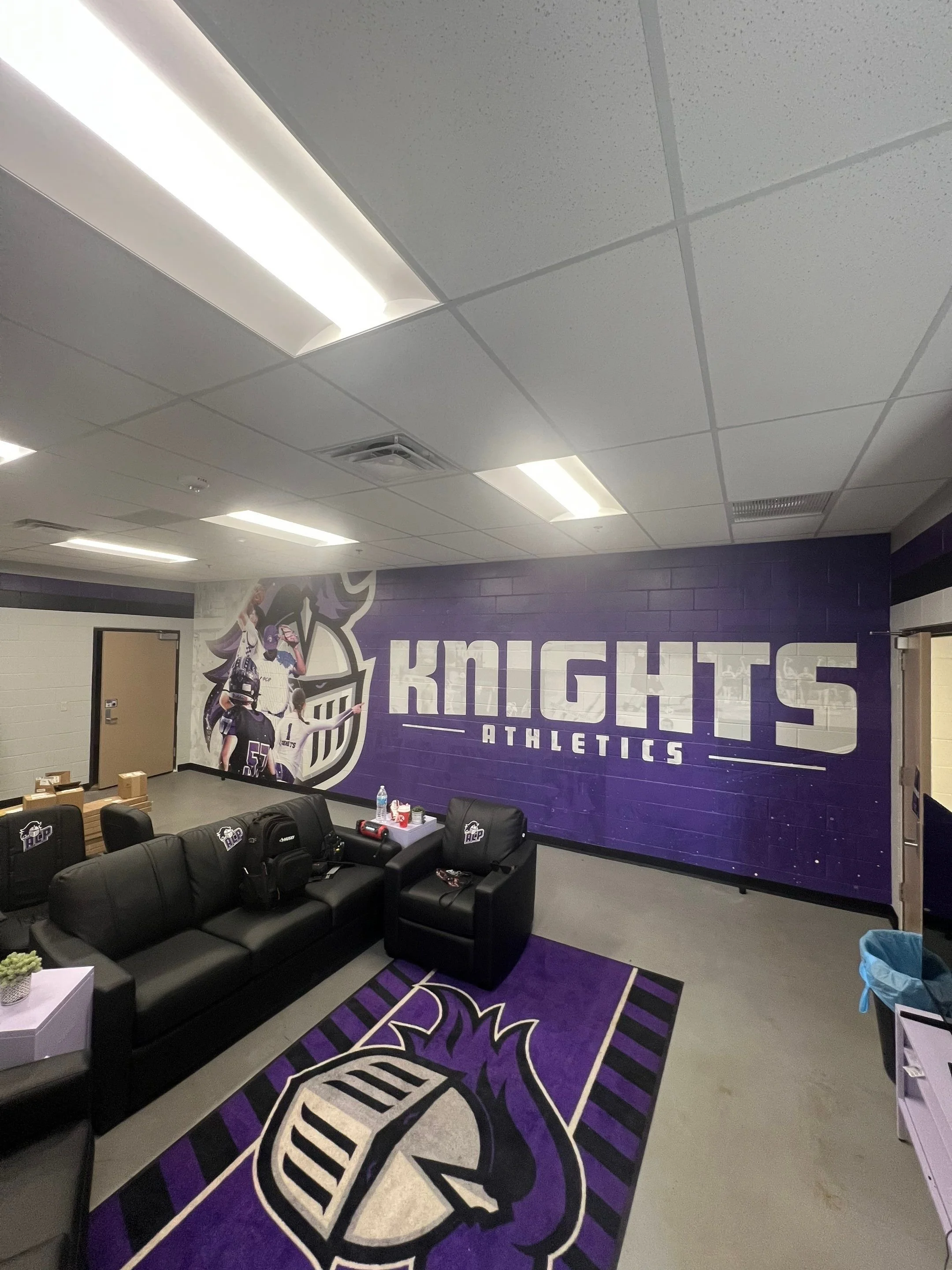 Locker room with black couches, purple and black team logo rug, and a large purple wall with white text reading 'KRUCHT ATHLETICS' along with a graphic of a rugby player and the team logo