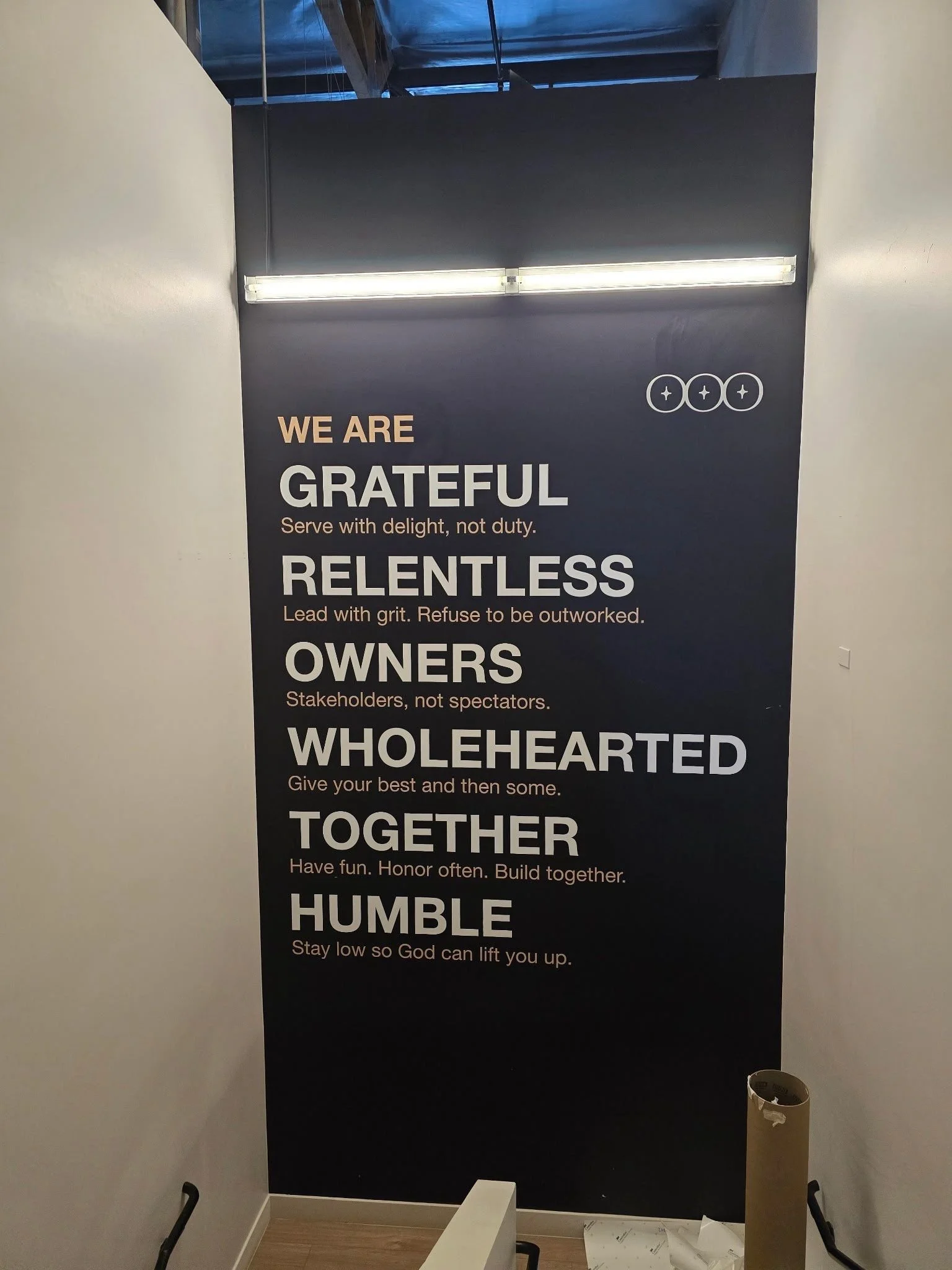 A black wall with motivational words and phrases in large white and tan letters, including 'WE ARE GRATEFUL,' 'RELENTLESS,' 'OWNERS,' 'WHOLEHEARTED,' 'TOGETHER,' and 'HUMBLE.' Additional smaller text is beneath each phrase. Part of a staircase, some 