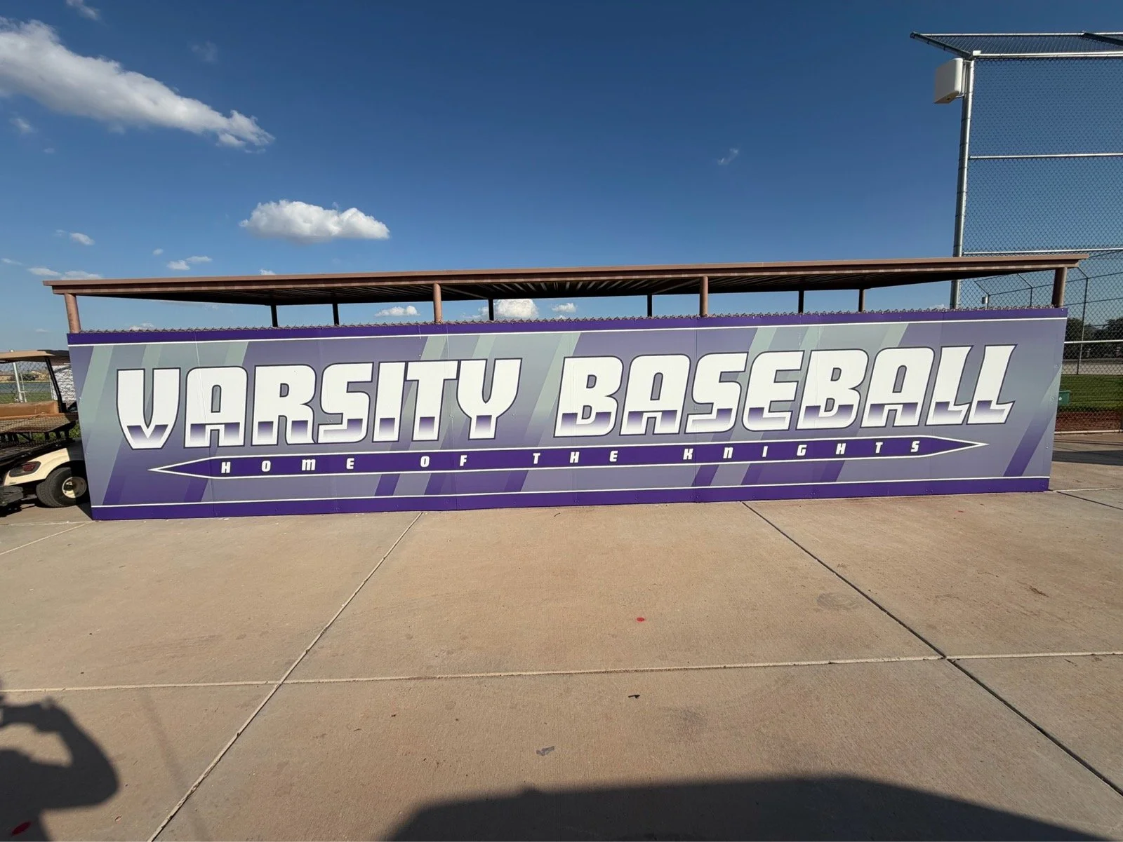 A large purple sign at a baseball field reads "VARSITY BASEBALL" with the subtitle "HOME OF THE KNIGHTS" and a light purple background with diagonal stripes.