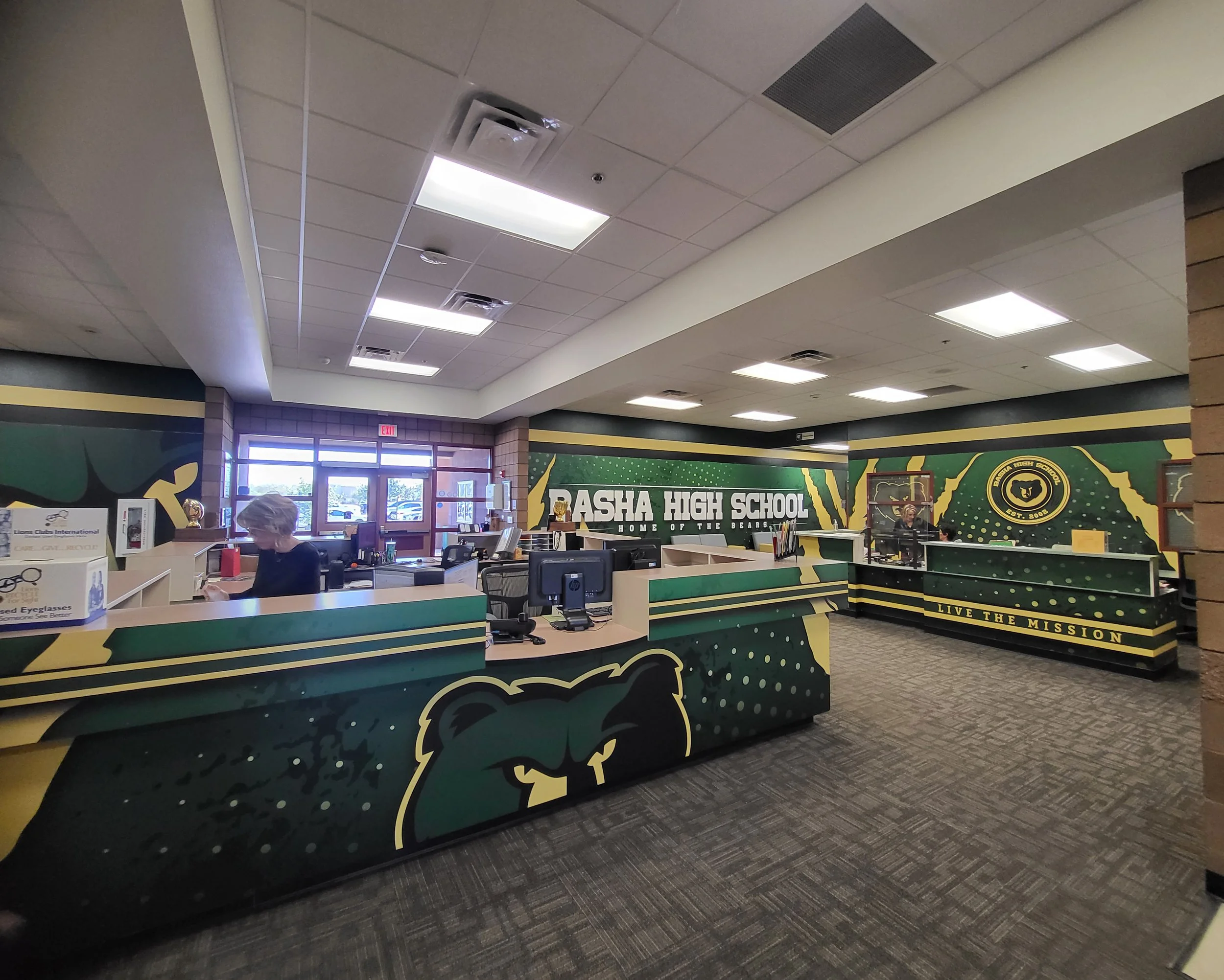 Inside a school office with a reception desk, a person behind the desk, and a large sign on the wall reading "Pasadena High School" with a bear mascot and the tagline "Home of the Bears."