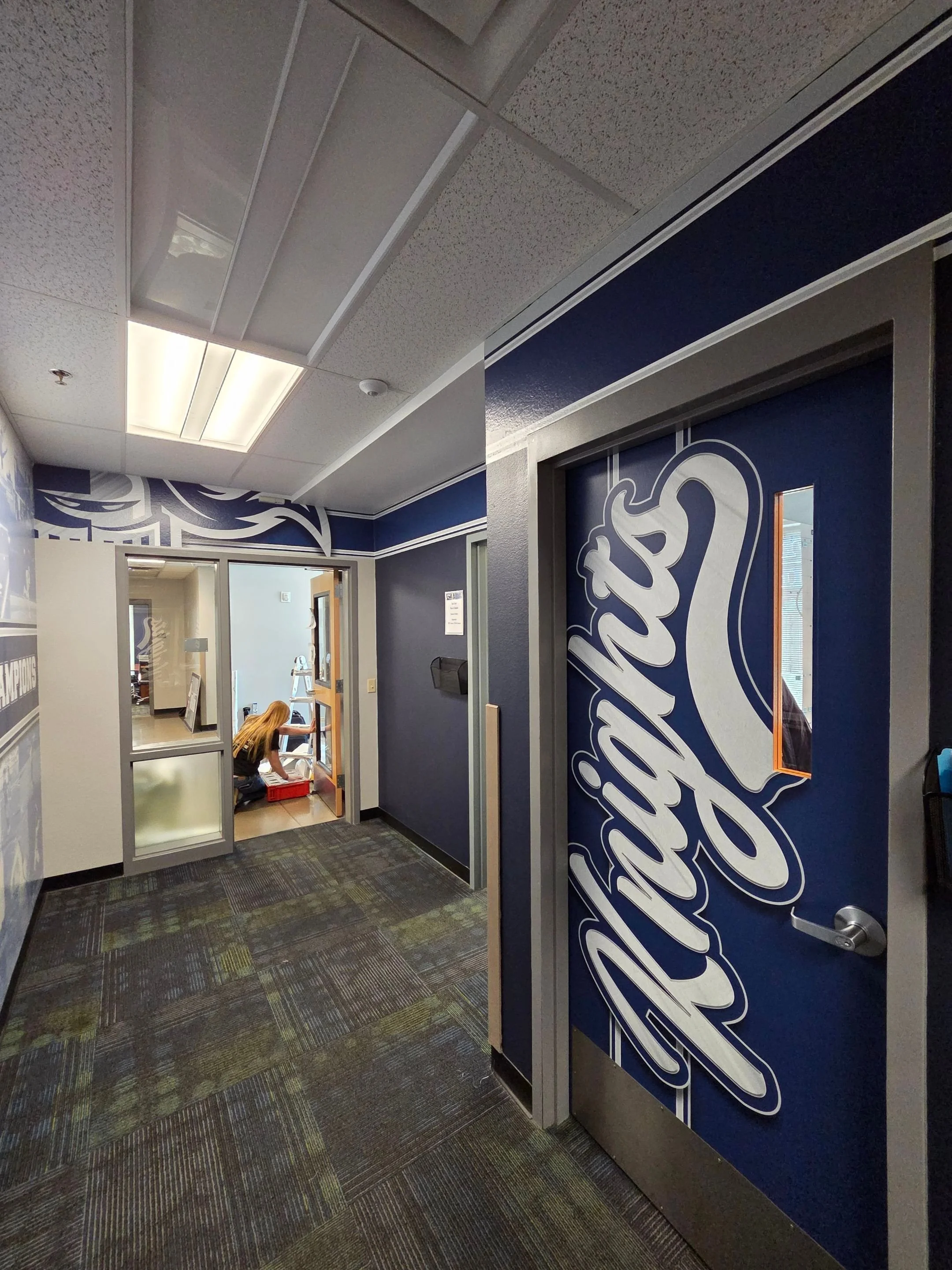 Office hallway with a door covered by a large blue sign with white cursive writing, and a woman working on a small ladder in a room through a glass door.