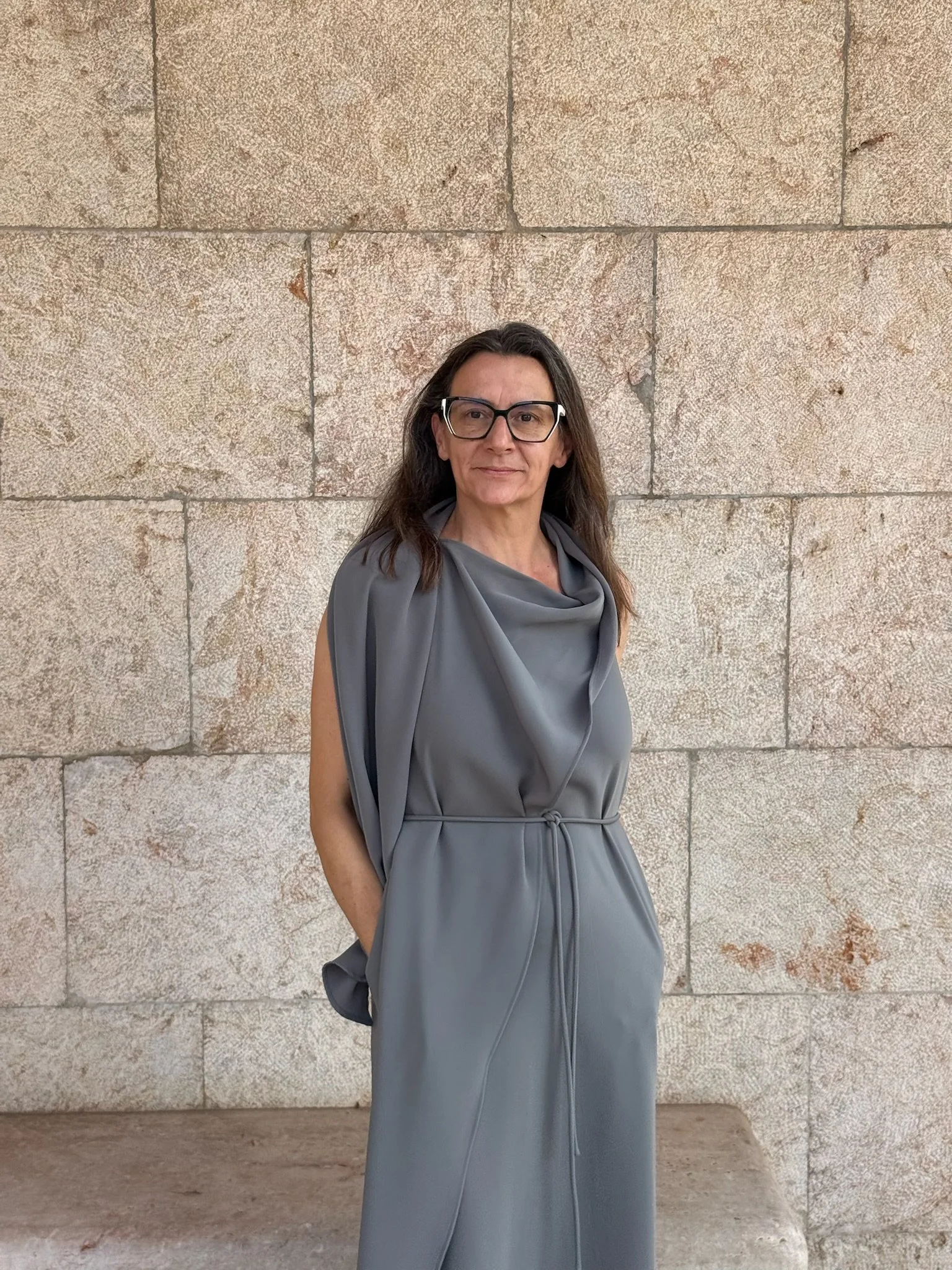 Mujer de cabello largo y gafas negras, vistiendo un vestido gris, de pie frente a una pared de piedra