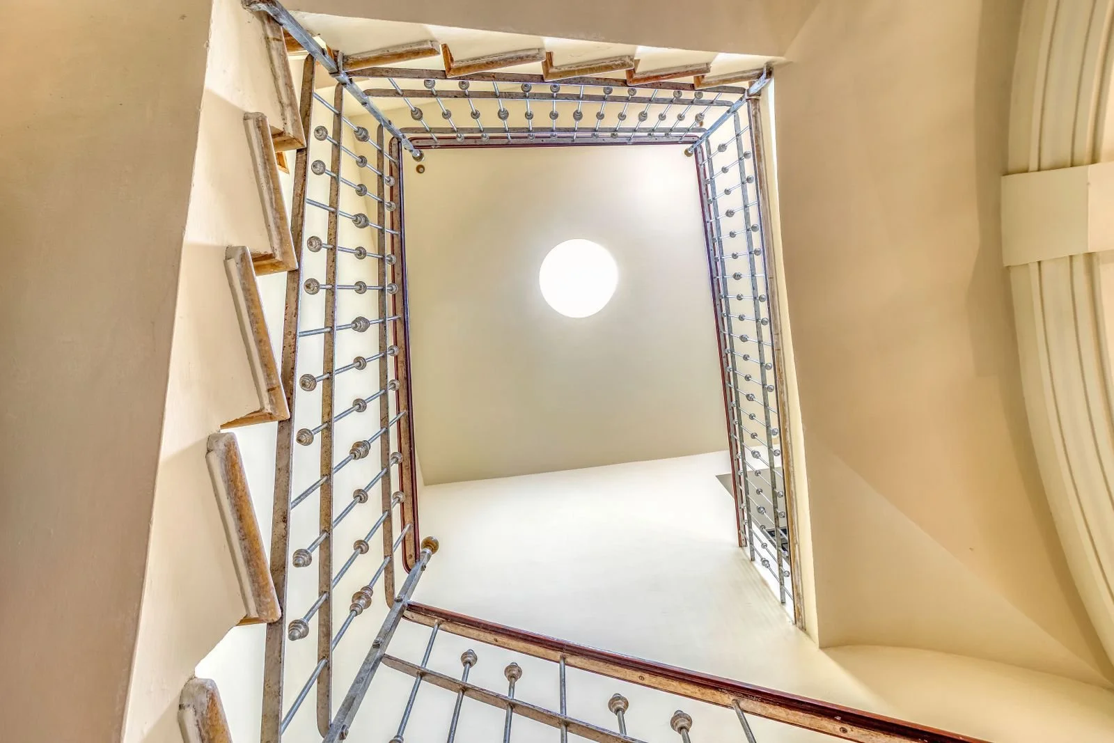 Vista hacia arriba de una escalera en un interior, con barandillas metálicas y luces en el techo, en un espacio con paredes beige y ventanas en los extremos.