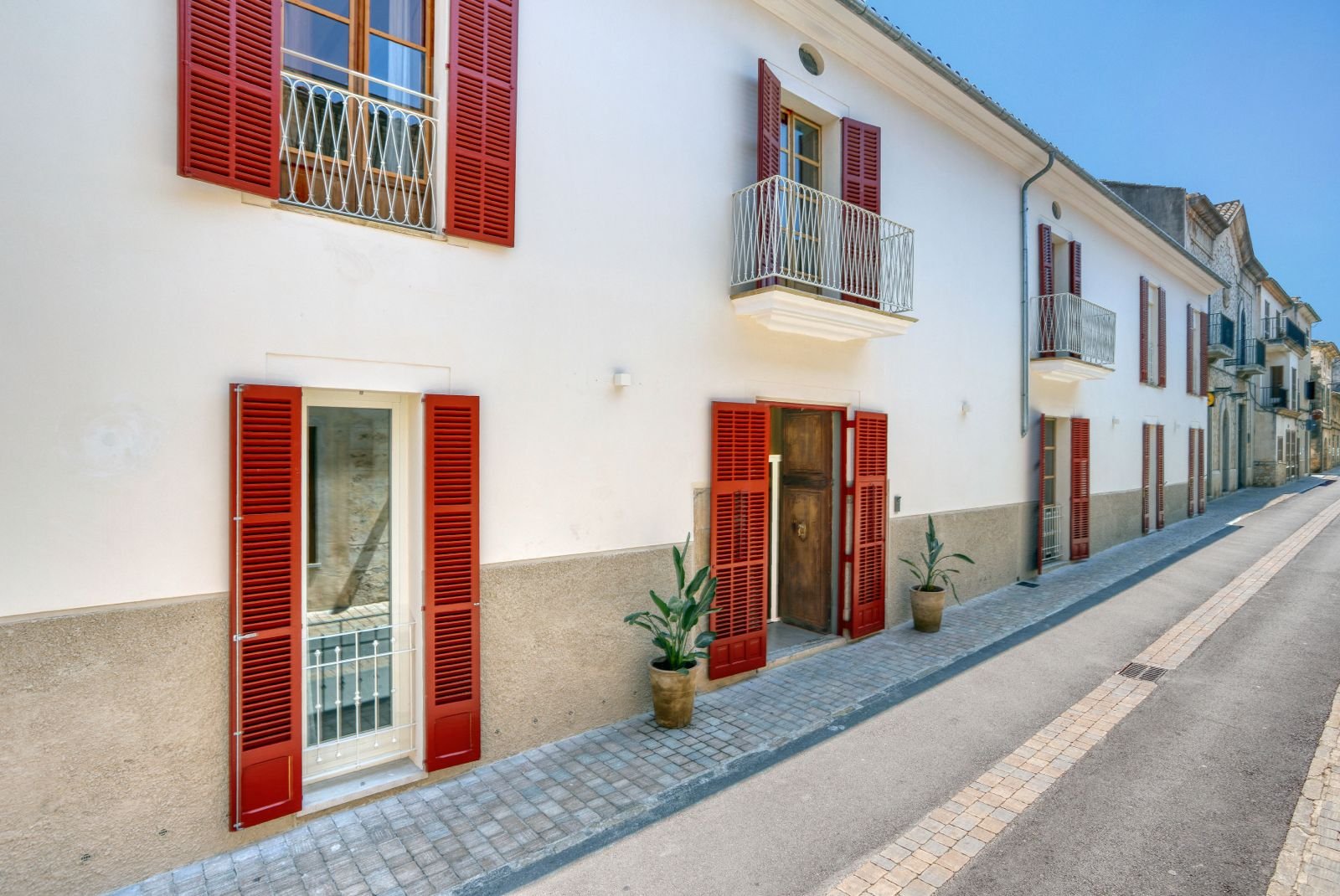 Edificio de color blanco con ventanas y puertas con contraventanas rojas en una calle pavimentada, con dos plantas y pequeñas terrazas con barandas blancas.