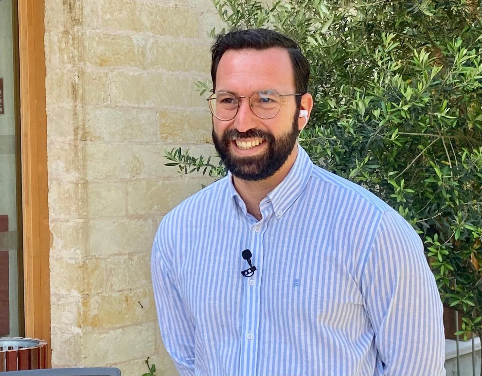 Hombre con barba, gafas, camisa de rayas azul y blanca, sonriendo, en un entorno con plantas y pared de piedra.