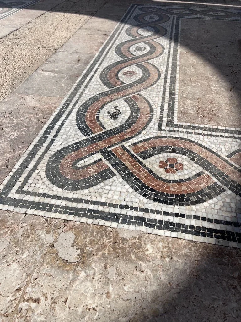 Mosaico decorativo en el suelo con patrones en espiral en tonos negros, blancos, y terracotas, en una zona de paso.