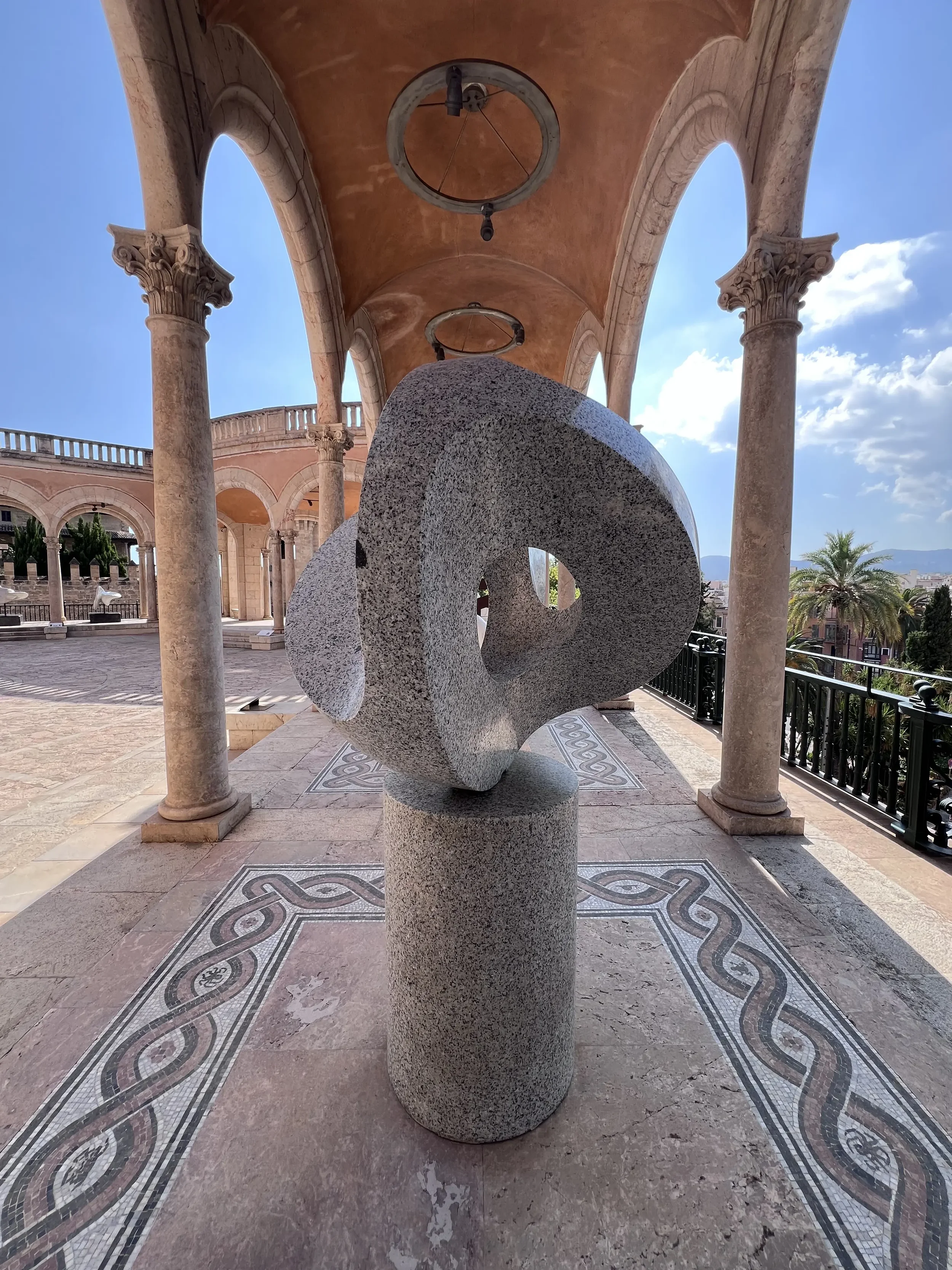 Escultura de piedra en forma espiral, ubicada en un lugar con columnas y techos con decoraciones arquitectónicas, cielo despejado y palmera en el fondo.