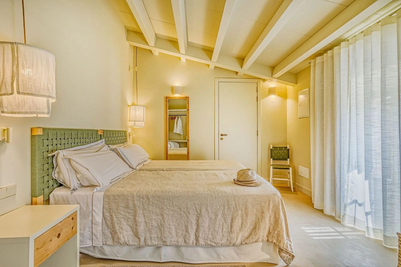 Habitación con cama doble, cortinas blancas, lámparas de pared, espejo, silla, y un sombrero en la cama.
