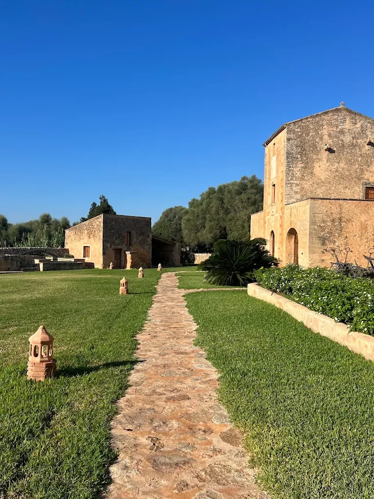 Camino de piedra que conduce a una casa antigua de piedra, rodeada de césped verde y plantas, con árboles grandes y un cielo despejado azul.