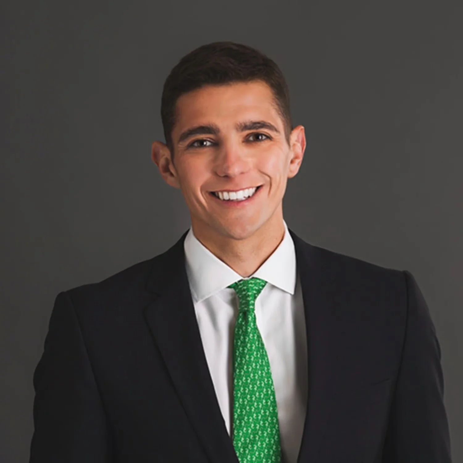 A young man in a black suit, white shirt, and green tie, smiling against a gray background.