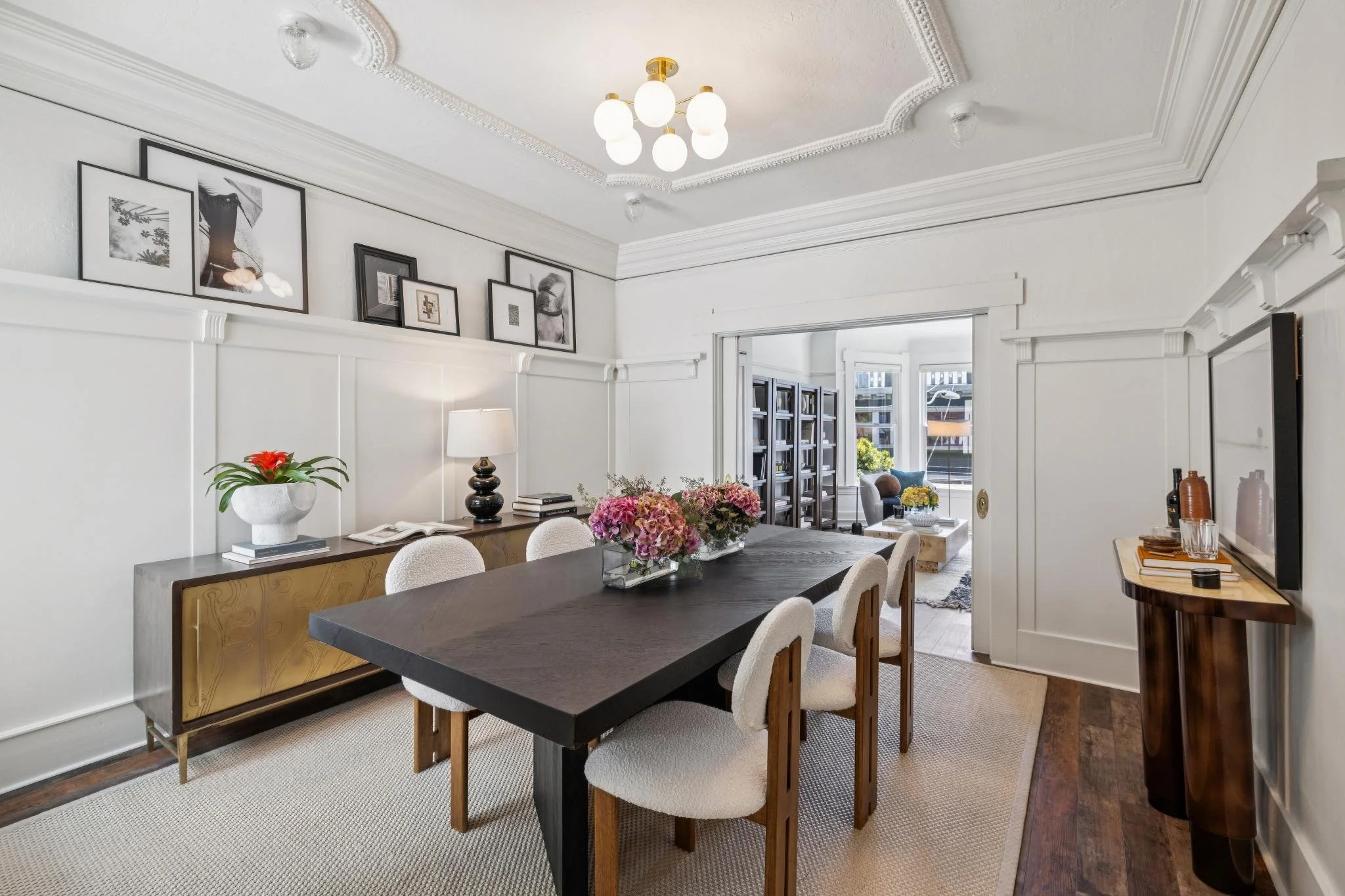 A dining room with a black table surrounded by white upholstered chairs, a wooden sideboard with a vase of pink flowers, framed black-and-white photos and artwork on the wall, a table lamp, and a view into a living area with big windows and additiona
