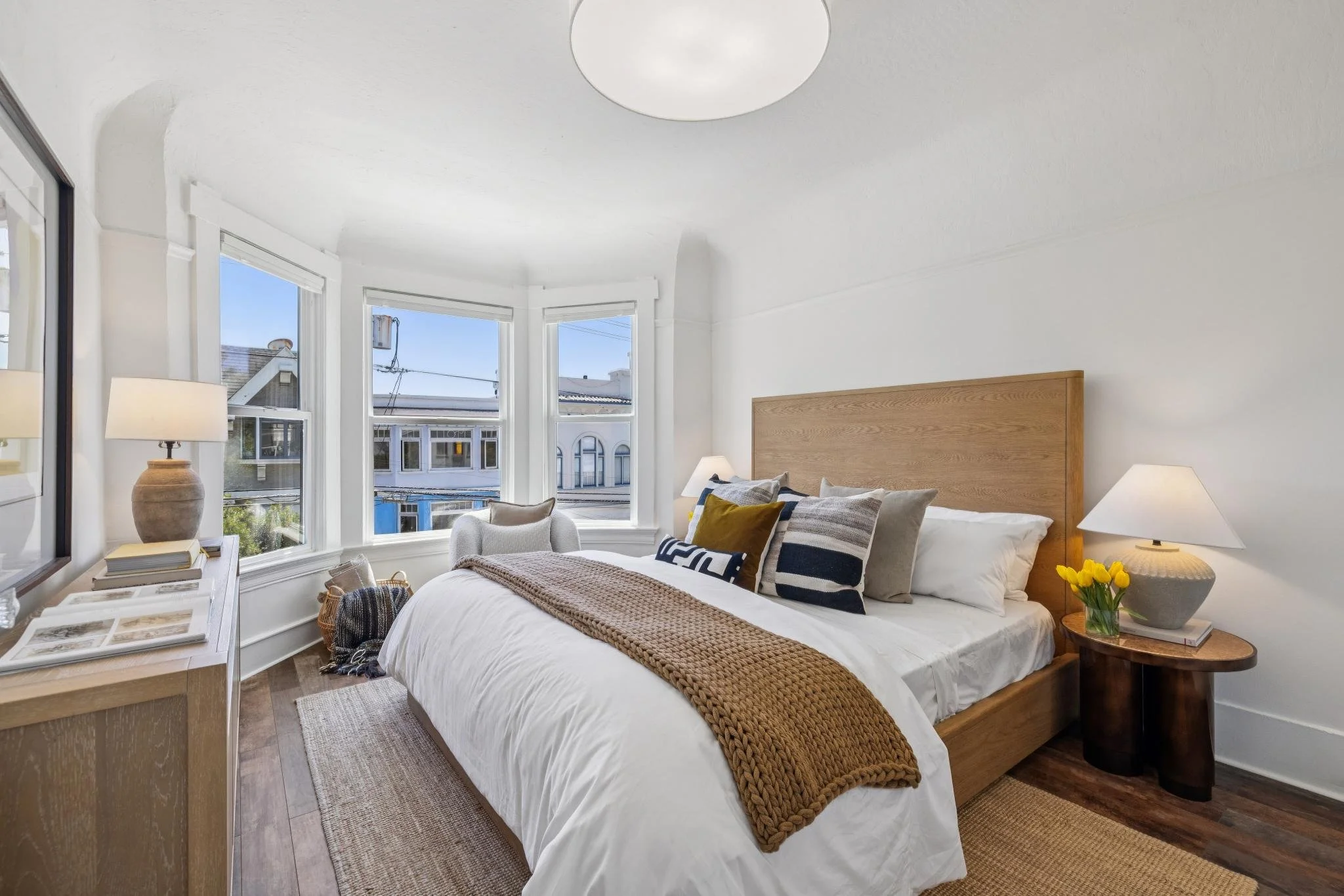 Bright bedroom with white walls and large bay windows showing neighboring houses and blue sky. The bed has white linens, striped pillows, and a chunky brown throw. Nightstand with a lamp, a vase of yellow flowers, and books. Wooden flooring with a wo
