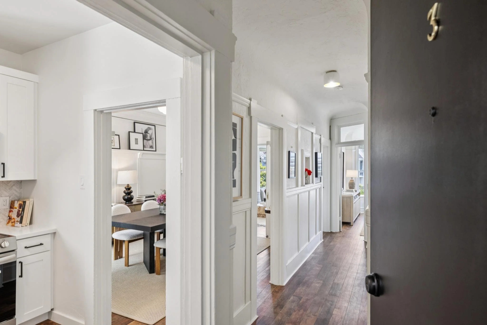 View of a bright, modern hallway in a house, with white walls, hardwood floors, and multiple doorways leading to different rooms. Natural light streams in through large windows at the end.
