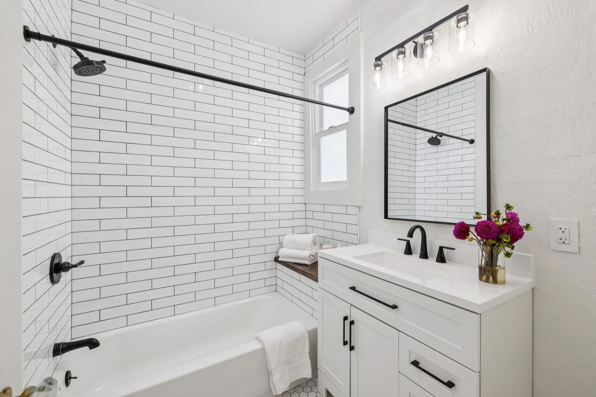 Bathroom with white subway tiles, a bathtub, a black shower fixture, a white vanity with black handles, a mirror, a window, a light fixture with four bulbs, and a vase of pink flowers.