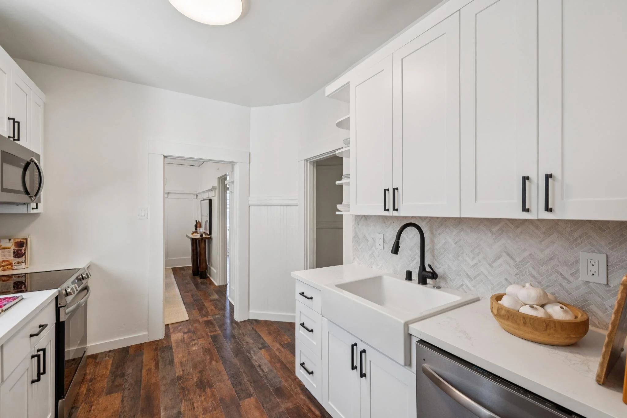 A modern, bright kitchen with white cabinets, black handles, a white countertop, and a white tile backsplash. There is a black faucet, a bowl of garlic on the counter, and a stainless steel microwave installed in the cabinets. The kitchen has dark wo