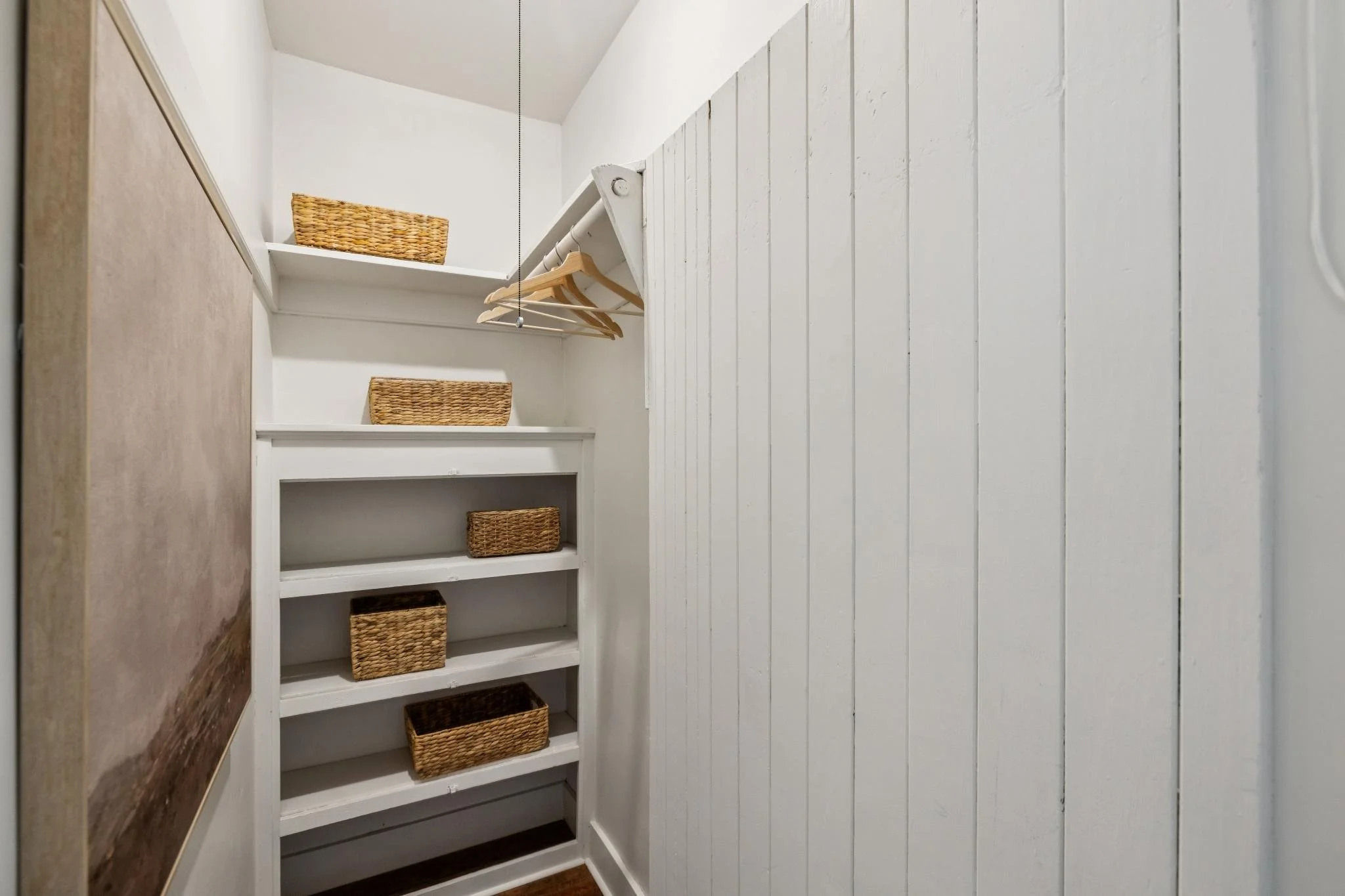 Small closet with white wooden shelves holding woven baskets and a hanging rod with wooden hangers.