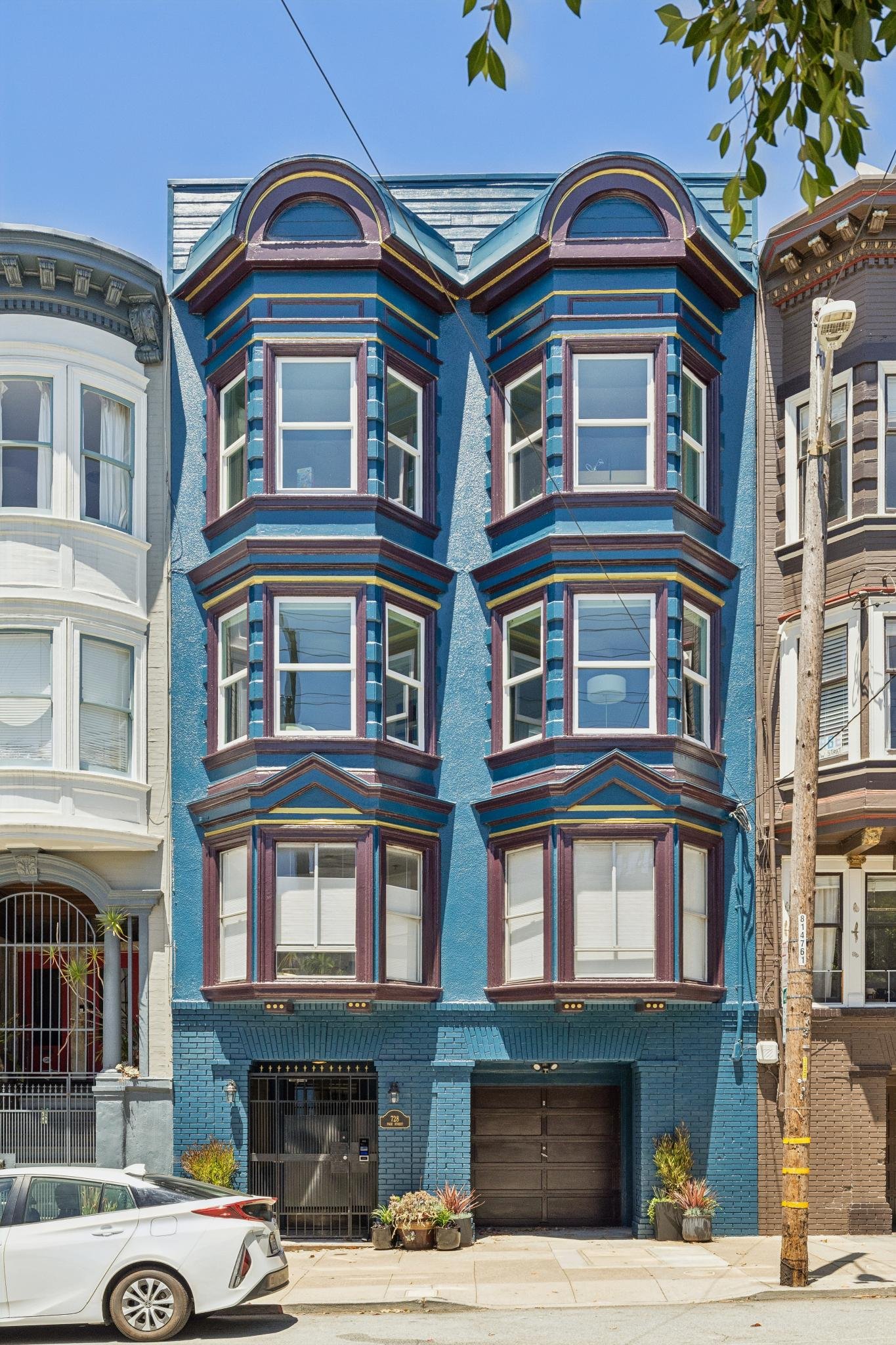 A three-story blue townhouse with prominent bay windows and detailed trim, situated between two other houses. A white car is parked in front, and potted plants are on the sidewalk. A utility pole with wires is on the right.