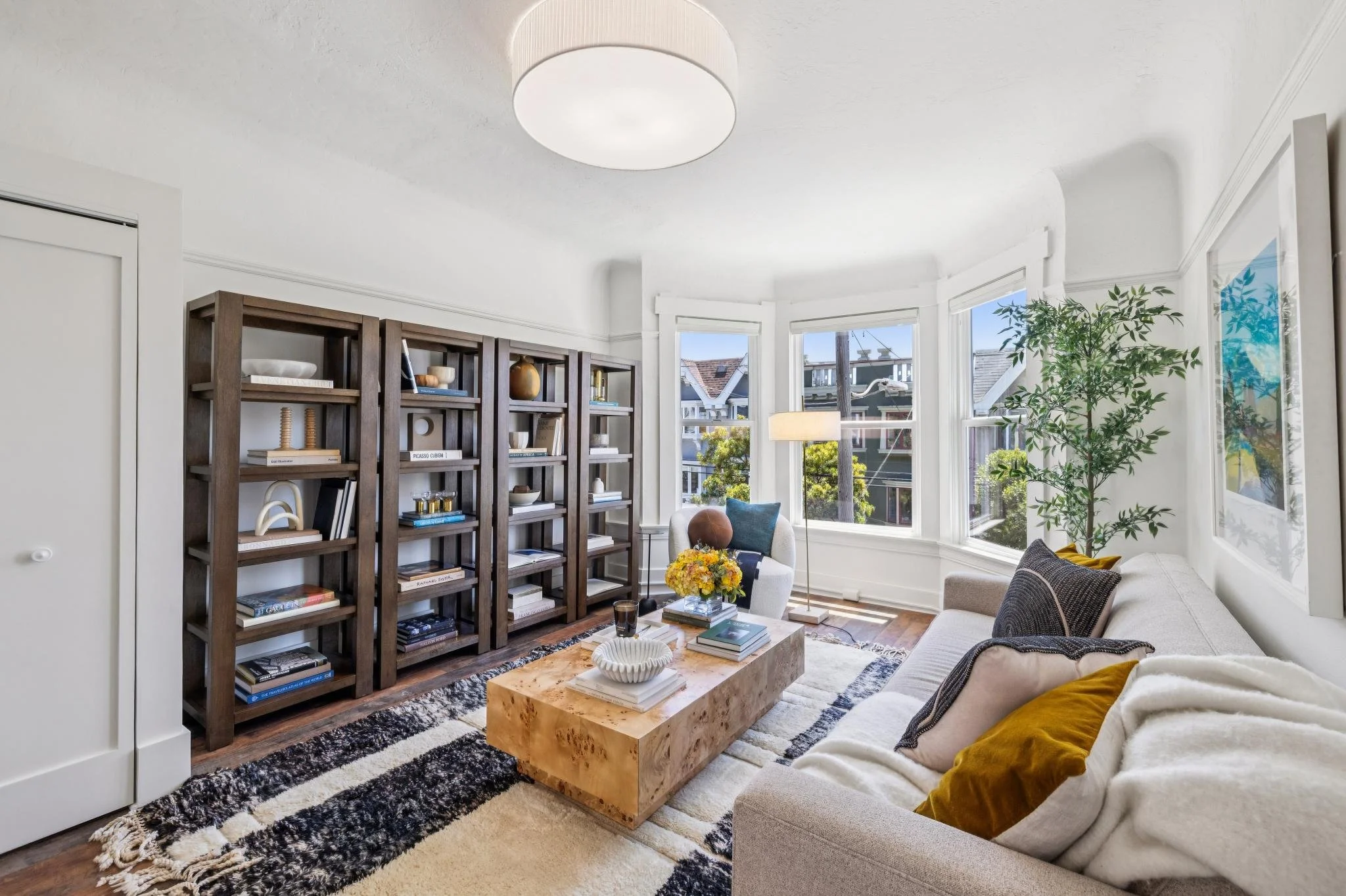A bright living room with large bay windows, a beige sofa with mustard and black pillows, a wooden bookcase, a zebra-patterned rug, a wooden coffee table with books and a white bowl, and a tall green plant. Sunny outdoor scene visible through the win