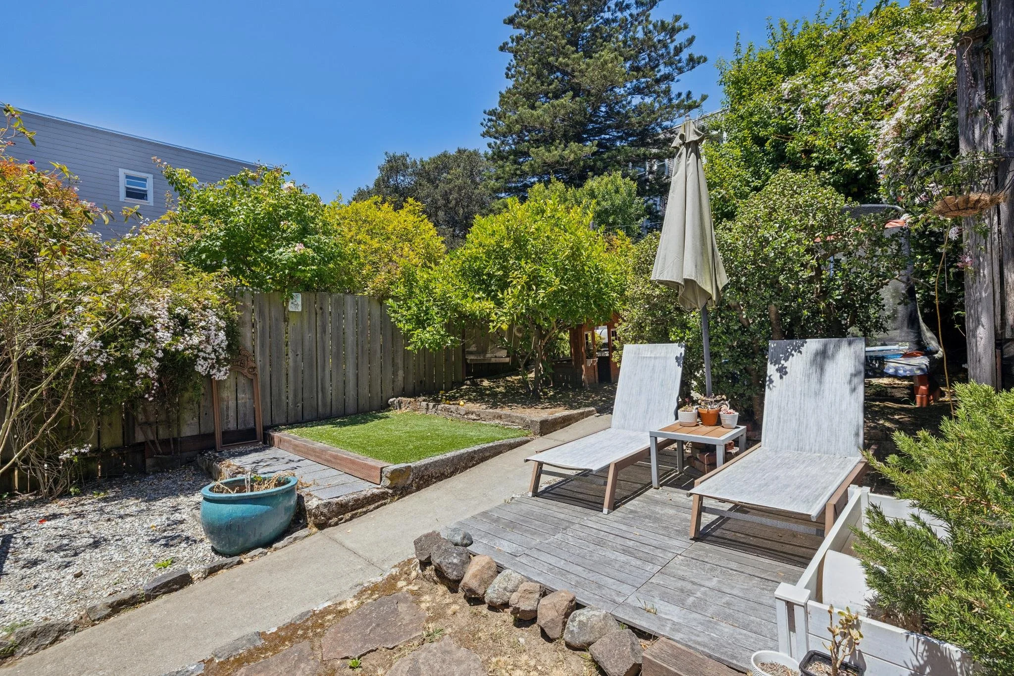 A backyard patio area with two lounge chairs, a small table with potted plants, a closed patio umbrella, and a well-maintained garden with trees, bushes, and flowers, enclosed by a wooden fence under a clear blue sky.