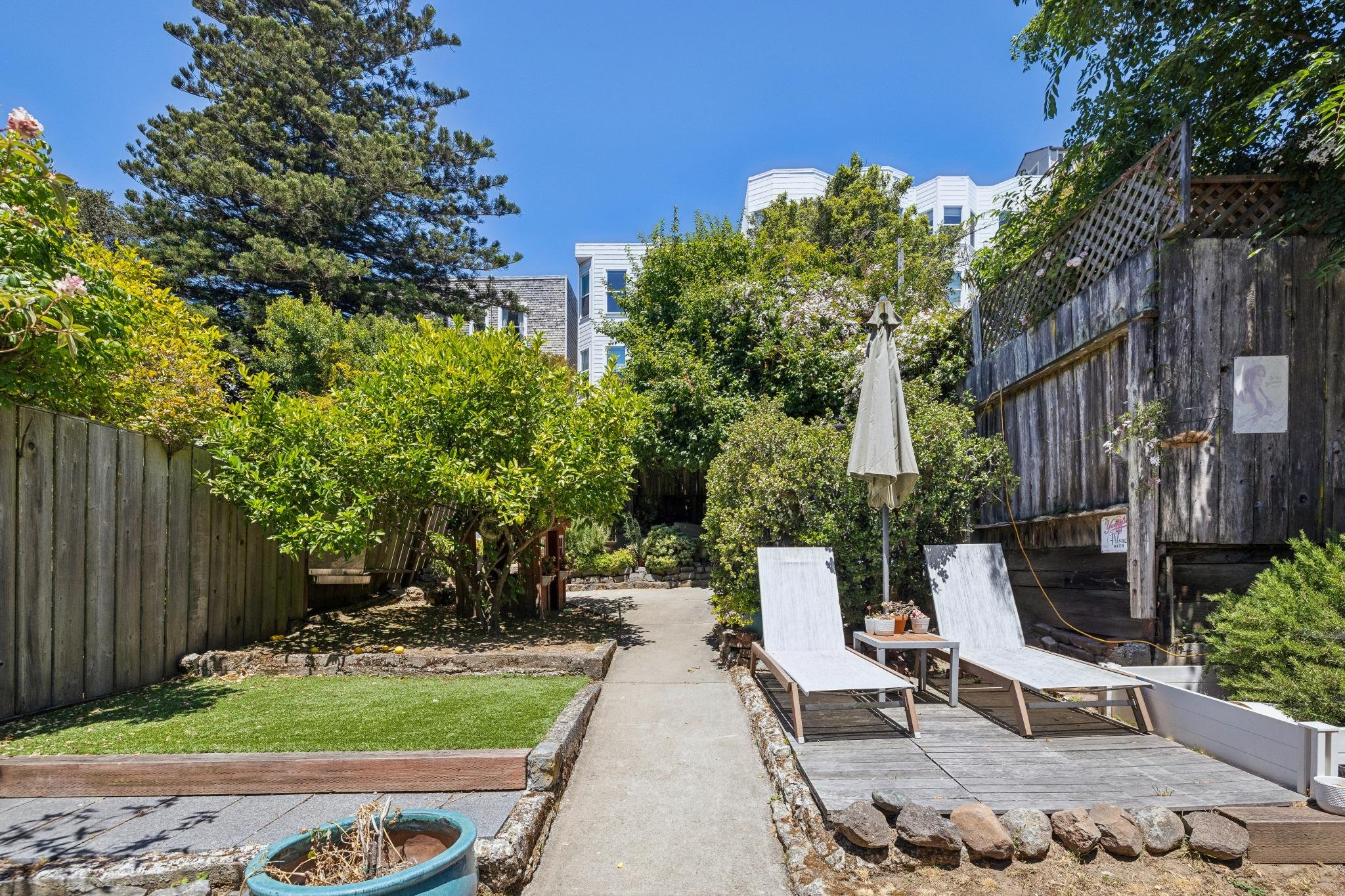 Backyard with trees, bushes, a wooden fence, and outdoor furniture including two white lounge chairs under a closed patio umbrella, with a blue sky above.