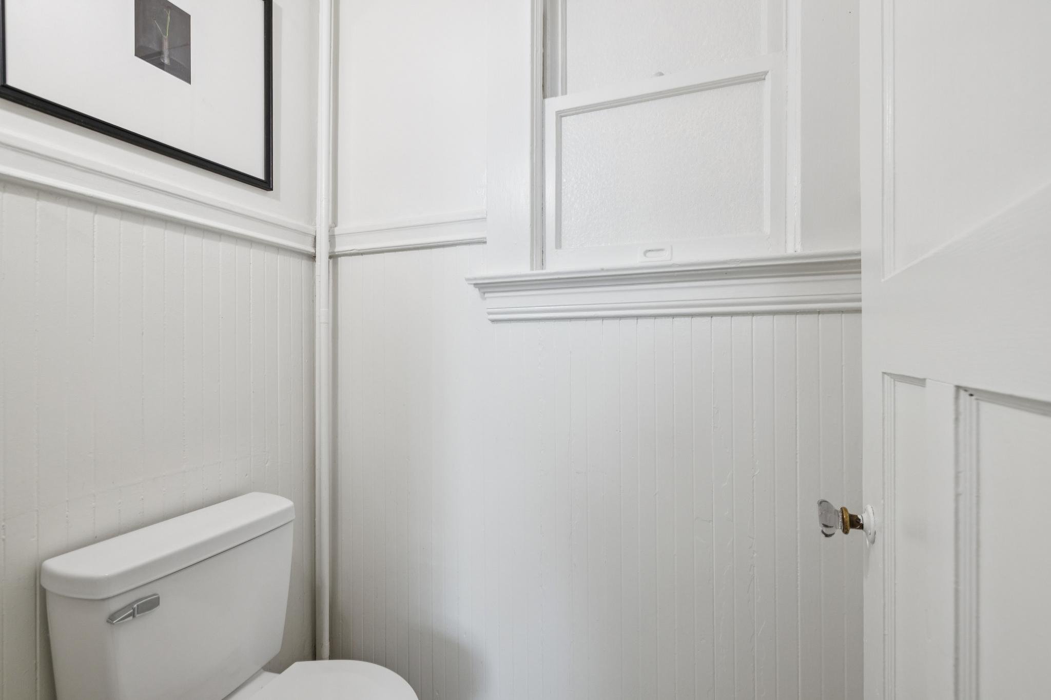 A small bathroom with white paneled walls, a toilet, a wall mirror, a small medicine cabinet, and a partially open door.