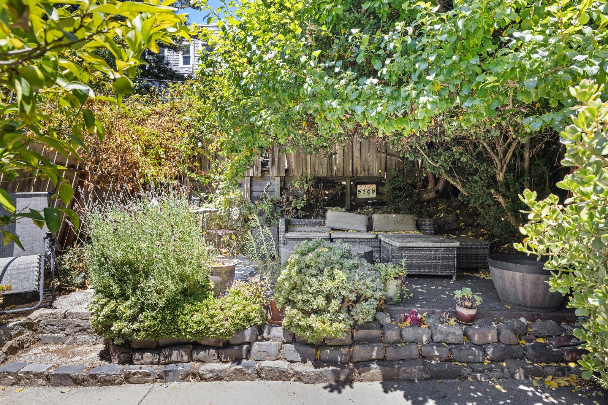 A backyard garden with potted plants, a seating area, and a wooden fence surrounded by lush green trees and shrubs.
