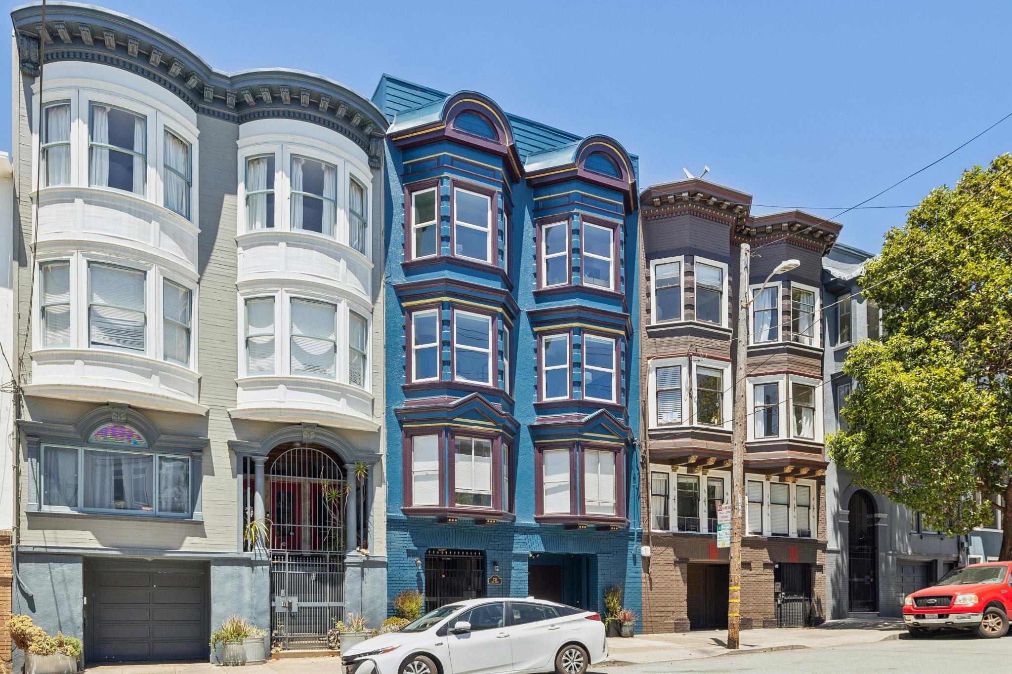 Colorful row of Victorian-style houses with ornate bay windows, painted in pastel shades of white, blue, and gray, under a clear blue sky with some trees and parked cars in front.