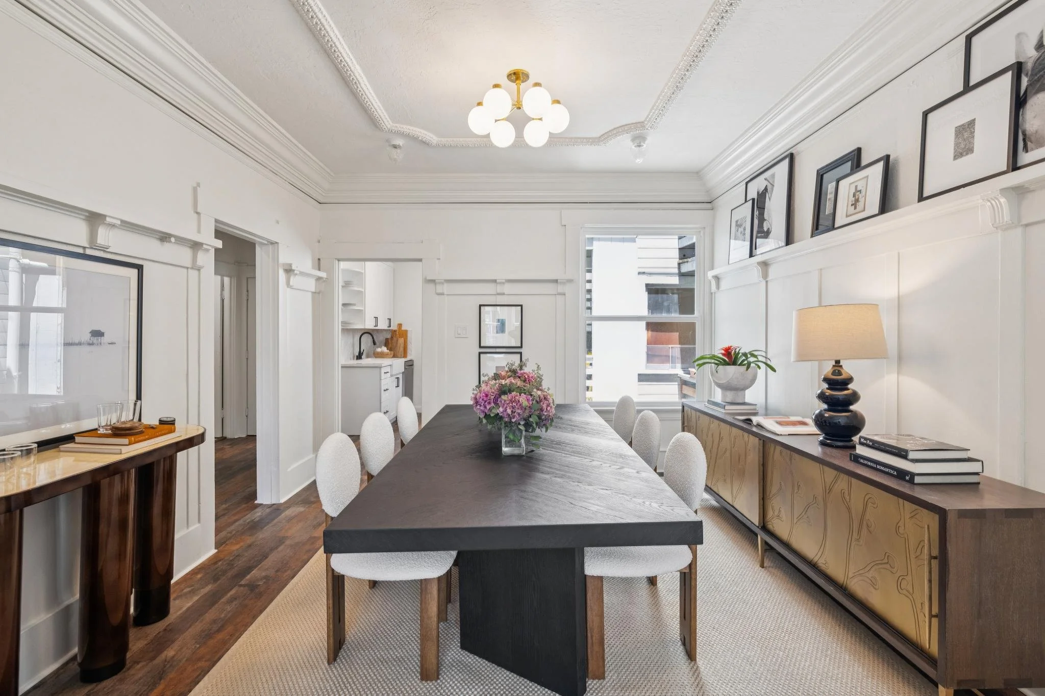 Modern dining room with a dark wood table, white upholstered chairs, a wooden sideboard with black accents, framed artwork on the wall, and a white ceiling with decorative molding and a gold chandelier. Large windows let in natural light, and there a