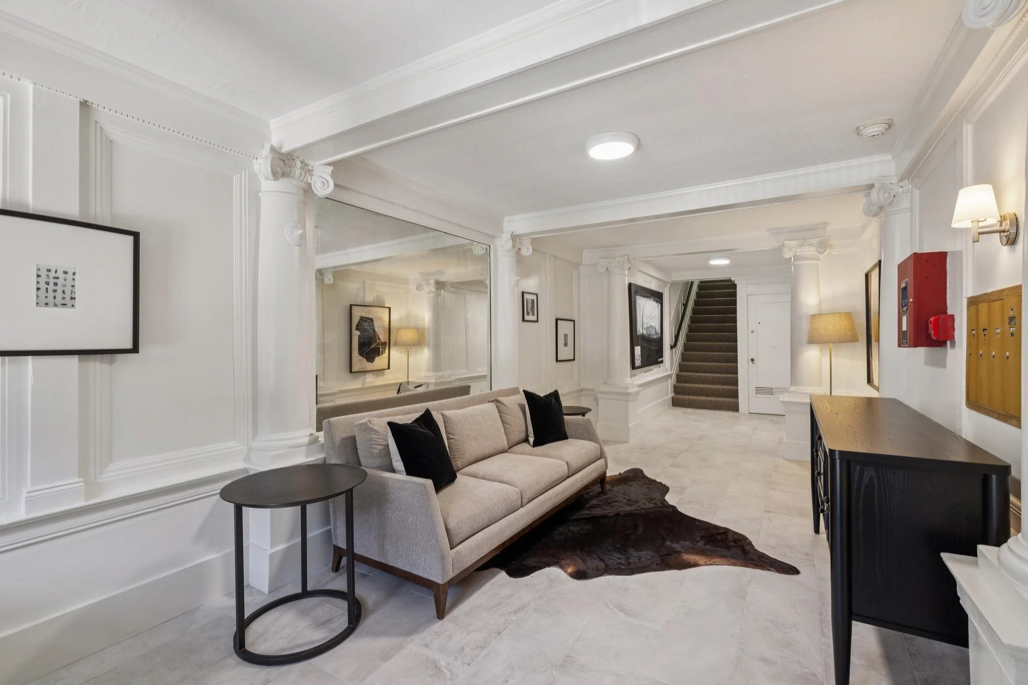 A modern living room with beige sofa, black accent pillows, side table, and wall art, featuring white walls, decorative columns, a staircase, and lighting fixtures.