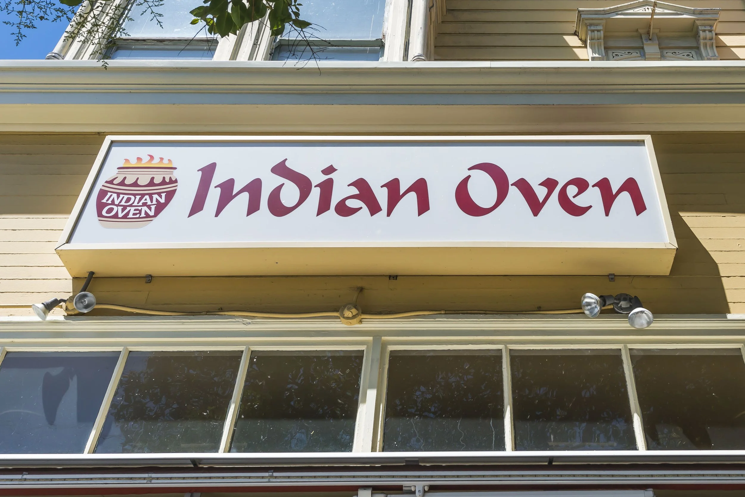 Sign for Indian Oven restaurant mounted on the exterior wall of a building, with windows above, two lights below, and visible blue sky.