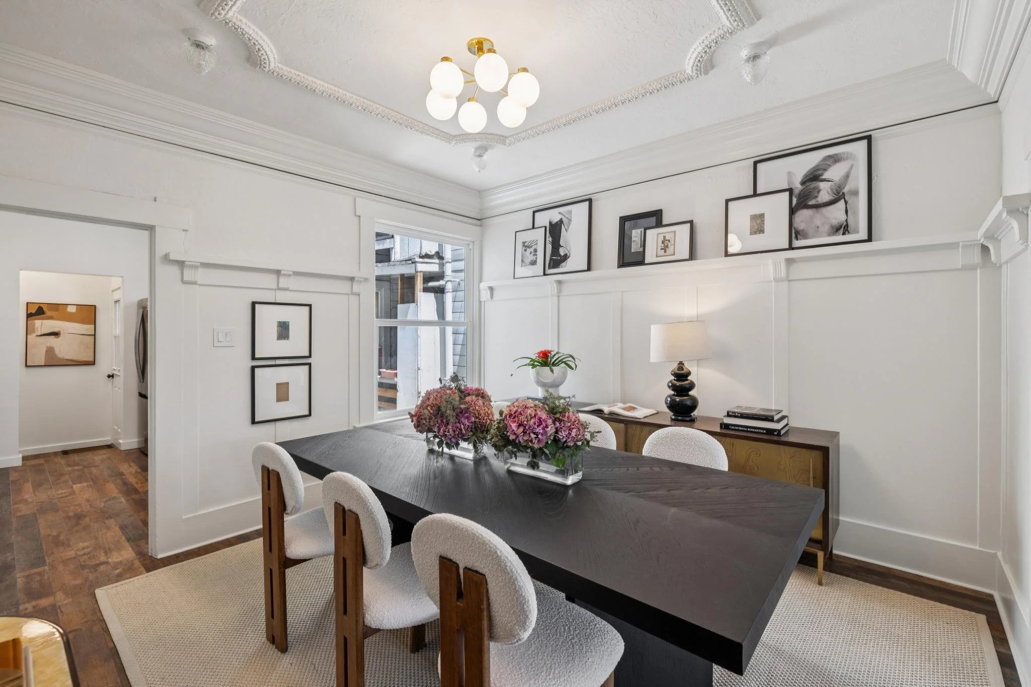 A dining room with a dark wooden table, white upholstered chairs, and a floral centerpiece of pink hydrangeas. The room has white walls, decorative crown molding, black and white artwork, a black sideboard, a black table lamp, and a large window with