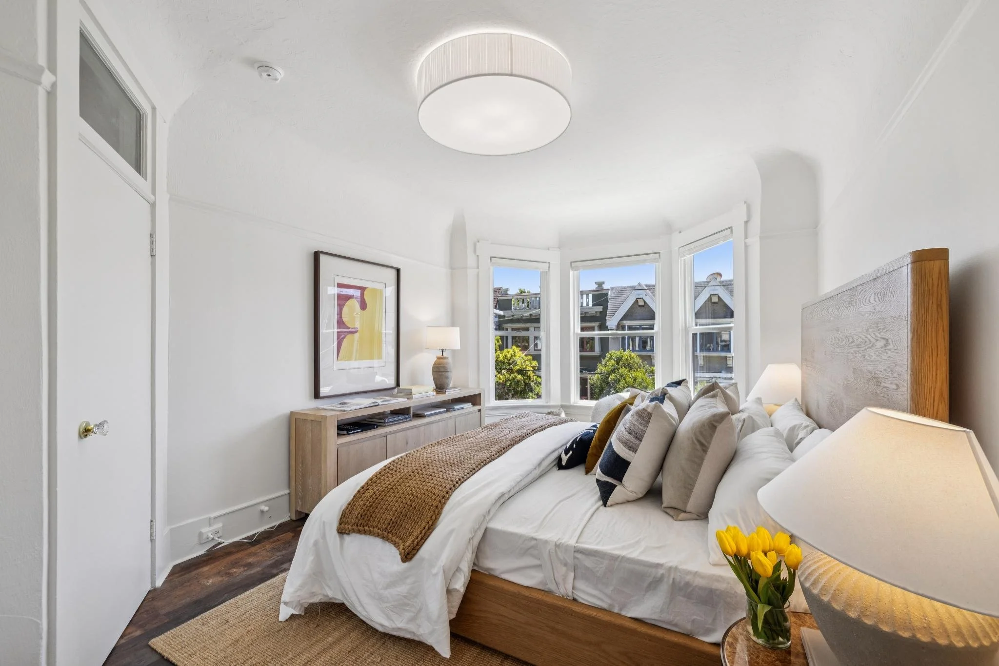 A cozy bedroom with a white bed, multiple pillows, and a wooden headboard. There is a bedside table with a lamp and a vase of yellow tulips. The room has a large bay window with views of neighboring houses and trees, and a framed abstract artwork on 