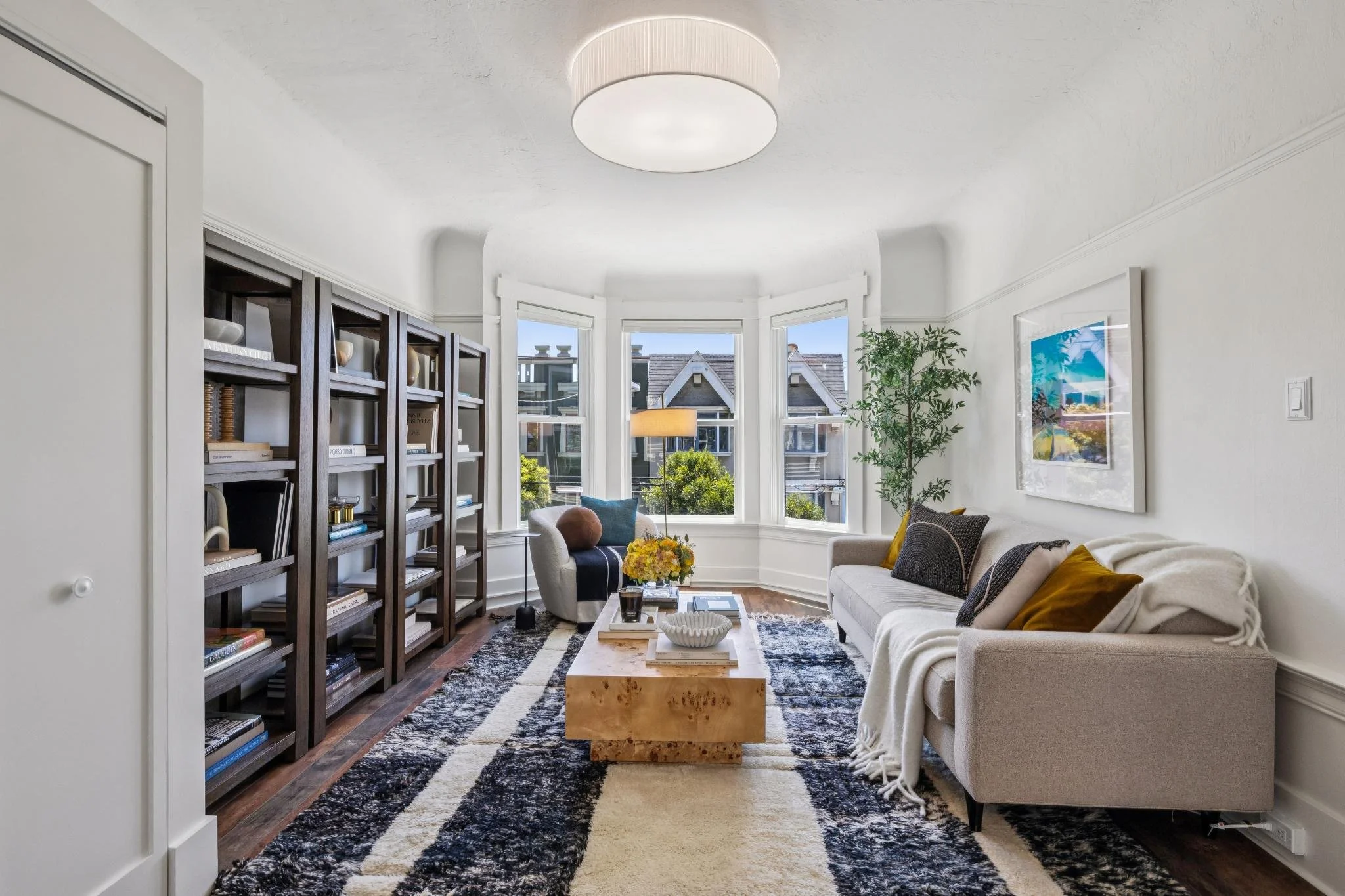Living room with large bay window, beige sofa with pillows, a white armchair, a black bookshelf filled with books and decorative items, a fireplace, a ceiling light, a coffee table with a vase of yellow flowers, a framed picture on the wall, and a mu