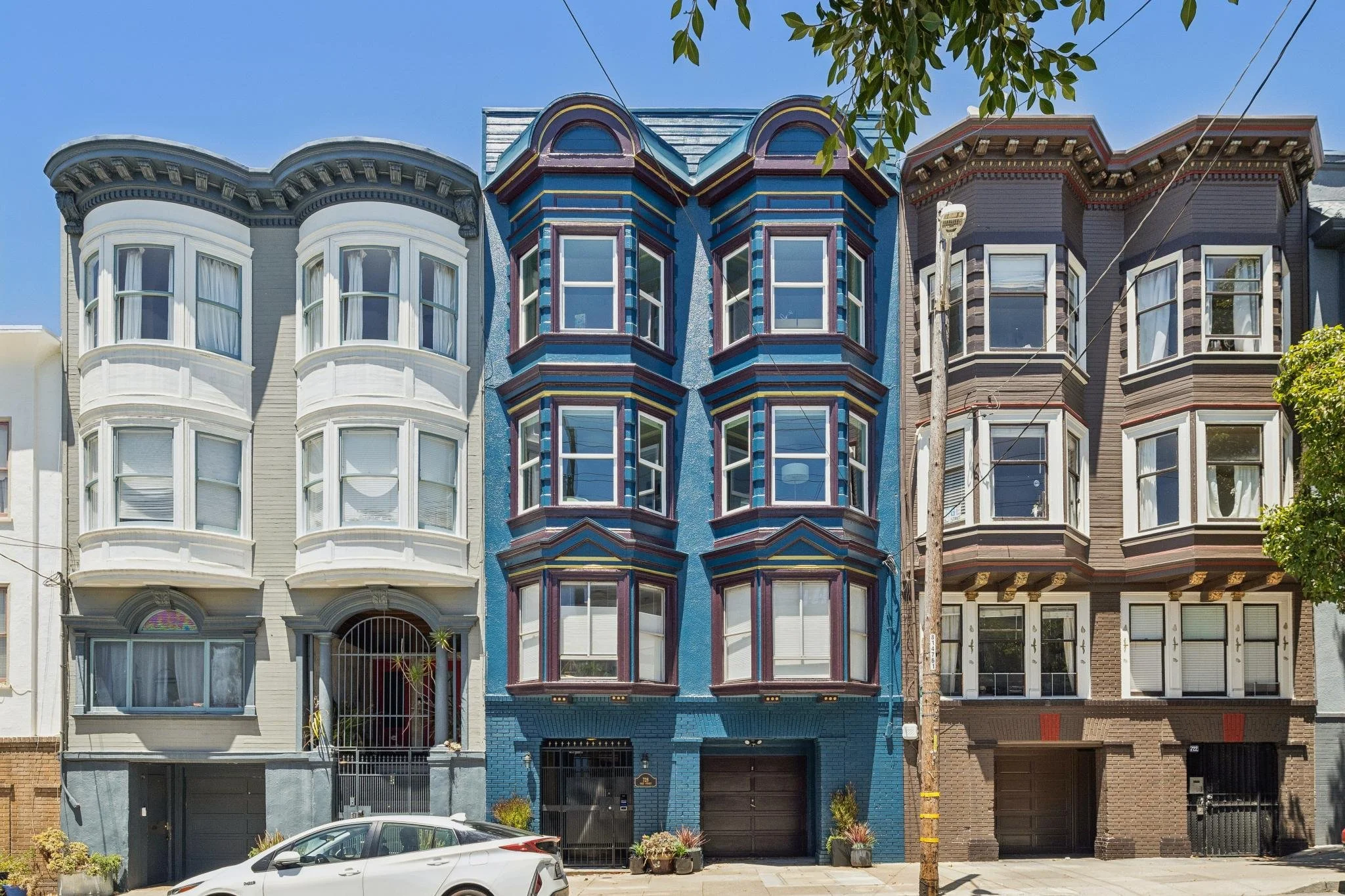 Three colorful Victorian-style houses in San Francisco with bay windows and ornate architectural details, lined up on a sunny city street with trees and a parked car in front.