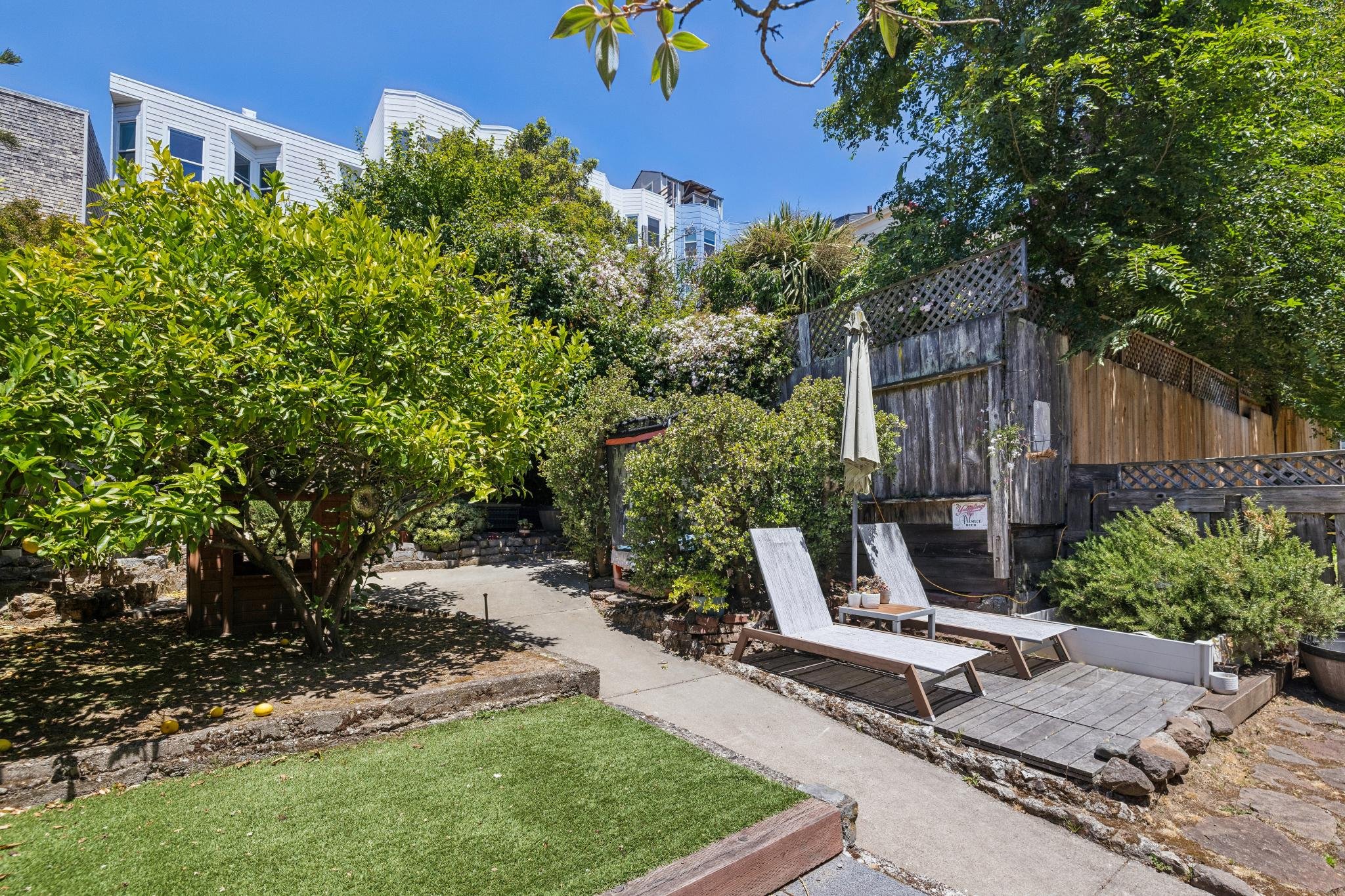 A backyard garden with green trees, bushes, and a patio area with chairs, a small deck, a wooden fence, and neighboring modern white buildings in the background under a clear blue sky.