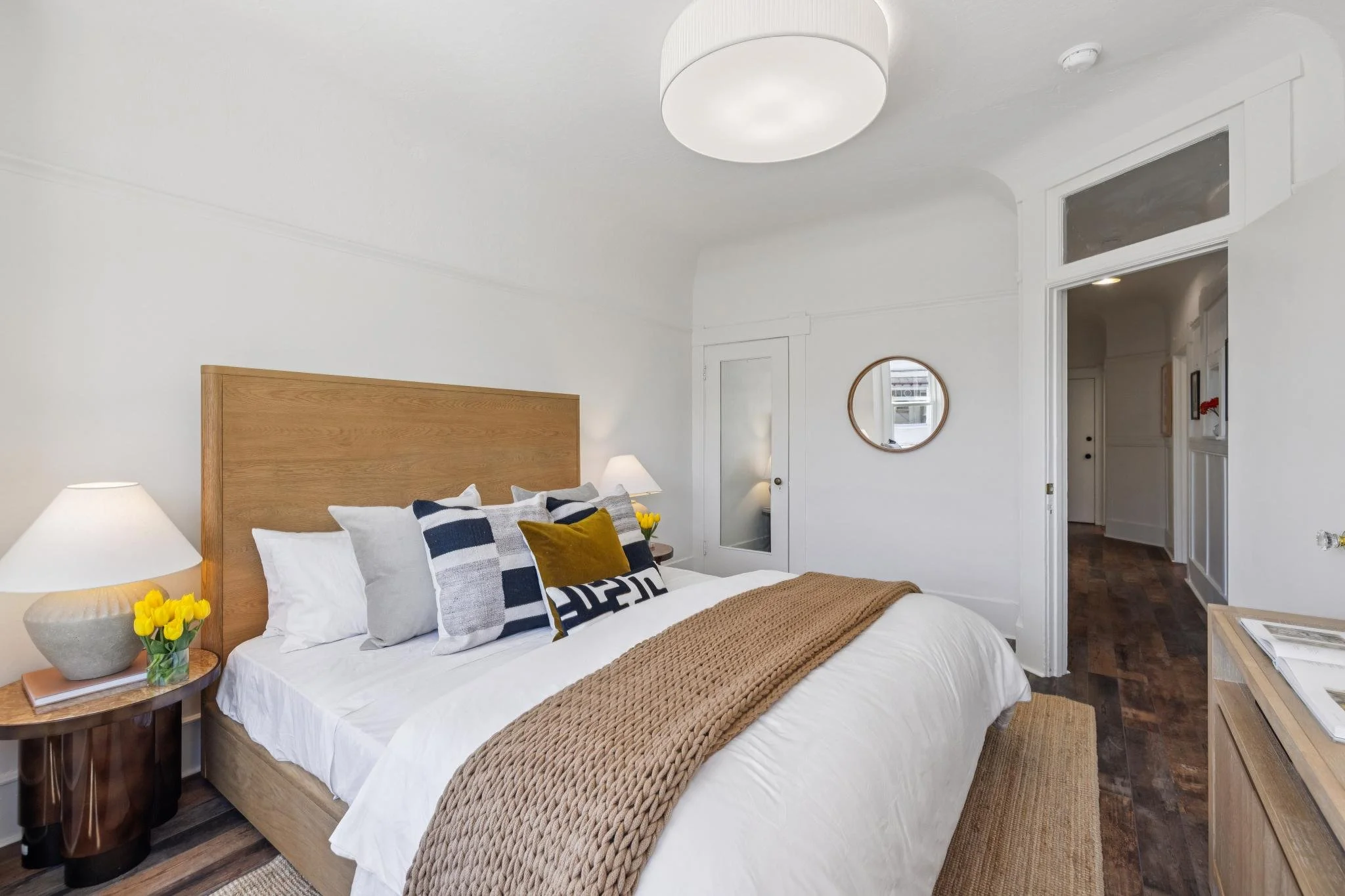 A bedroom with white walls, a wooden bed frame, and a nightstand with a lamp, a vase of yellow flowers, and books. The bed has white duvet, pillows, and a brown blanket. There is a mirror on the wall and a doorway leading to a hallway.