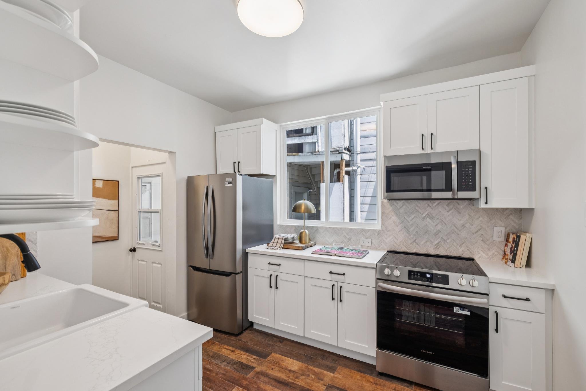 A modern kitchen with white cabinets, stainless steel refrigerator, microwave, and oven. There is a window above the counter with natural light, a gold desk lamp, and a wooden kitchen island with a white countertop.