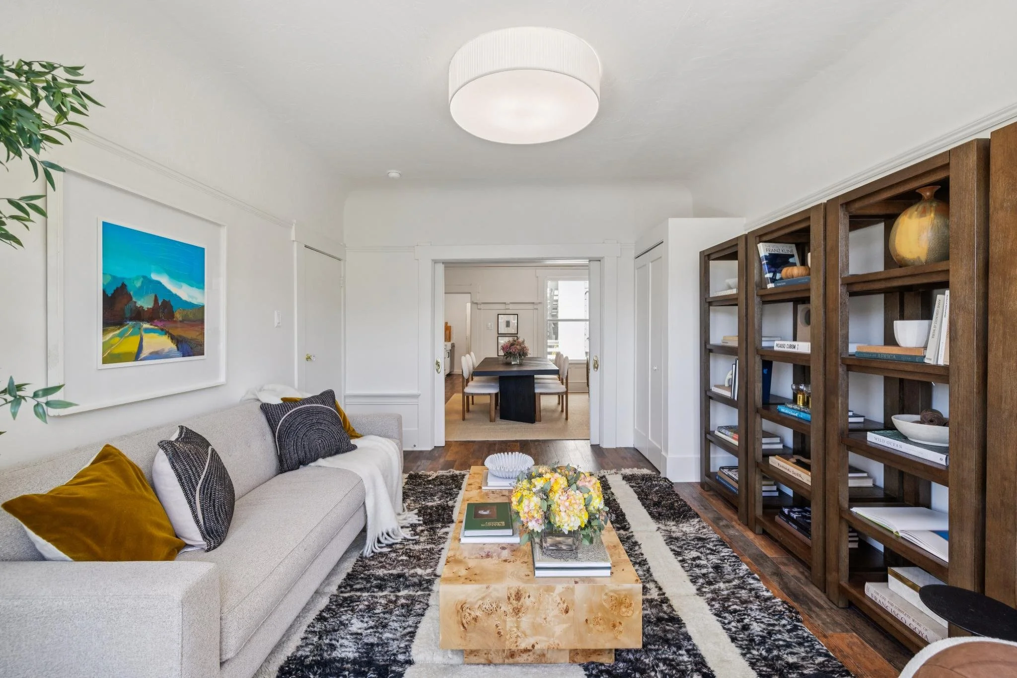 A living room with a gray sofa decorated with pillows, a wooden coffee table with a floral centerpiece and books, a patterned black and white rug, a bookshelf with decorative items, a colorful landscape painting on the wall, and a view into a dining 