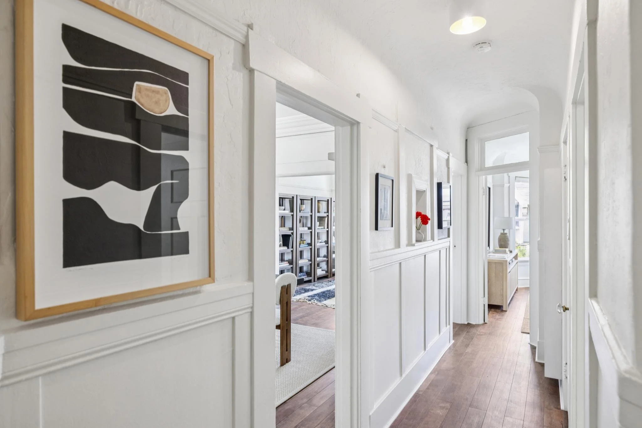 Bright hallway with white walls and large windows, decorated with framed artwork, bookshelf, vase with red flowers, and a table lamp.
