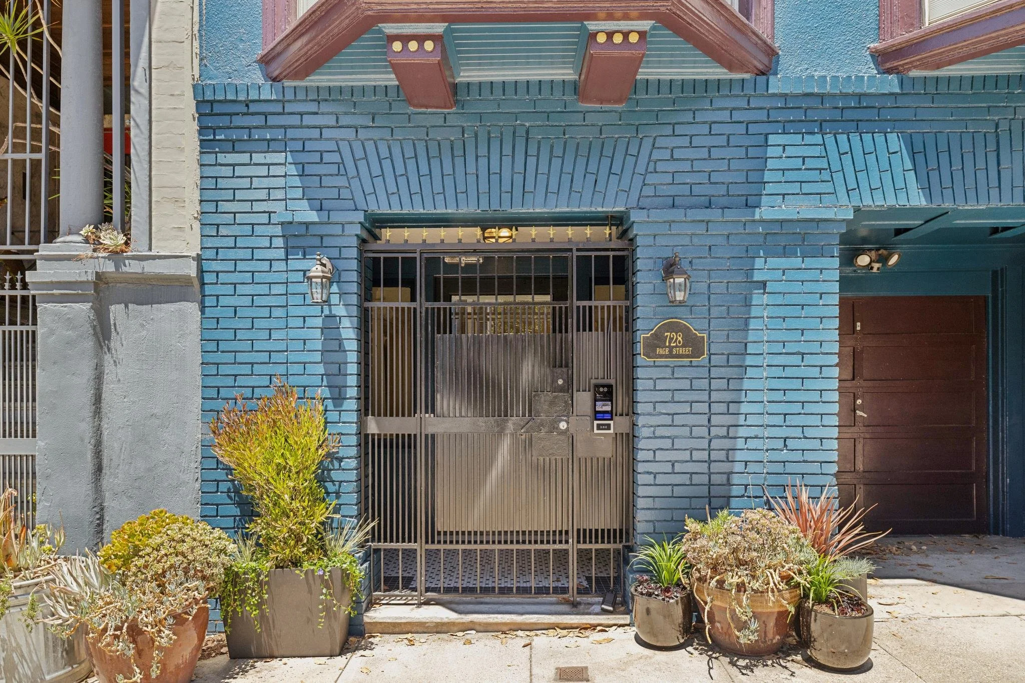 A blue brick building with brown window trim and a black gate at the entrance. Potted plants with succulents and greenery line the front of the building. The building has a small sign that reads '728 PAGE STREET' near the gate.