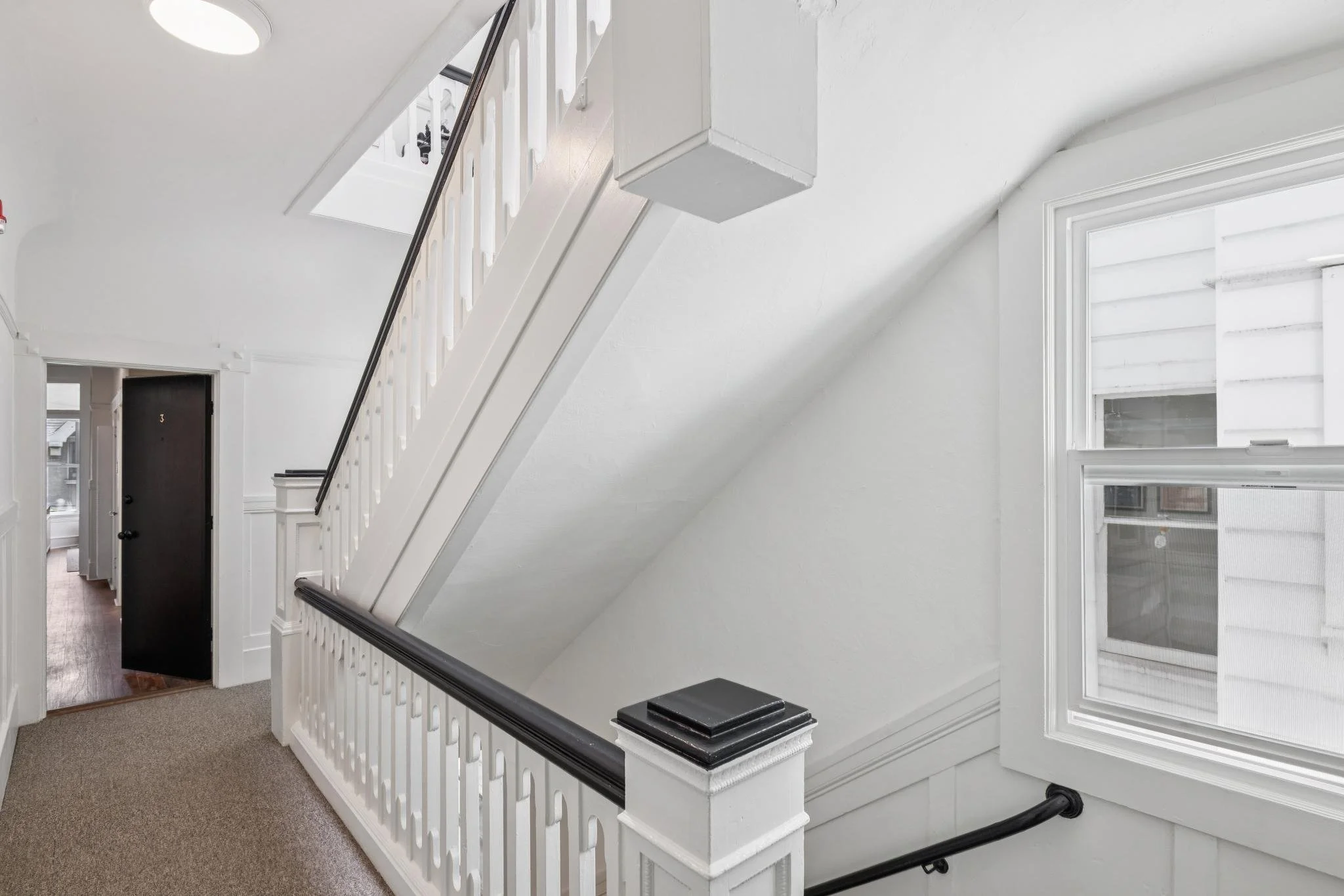Interior view of a house showing a staircase with white balusters and a black handrail, a window on the right side, and a hallway with a door in the background. The walls are painted white, and the floor is carpeted.